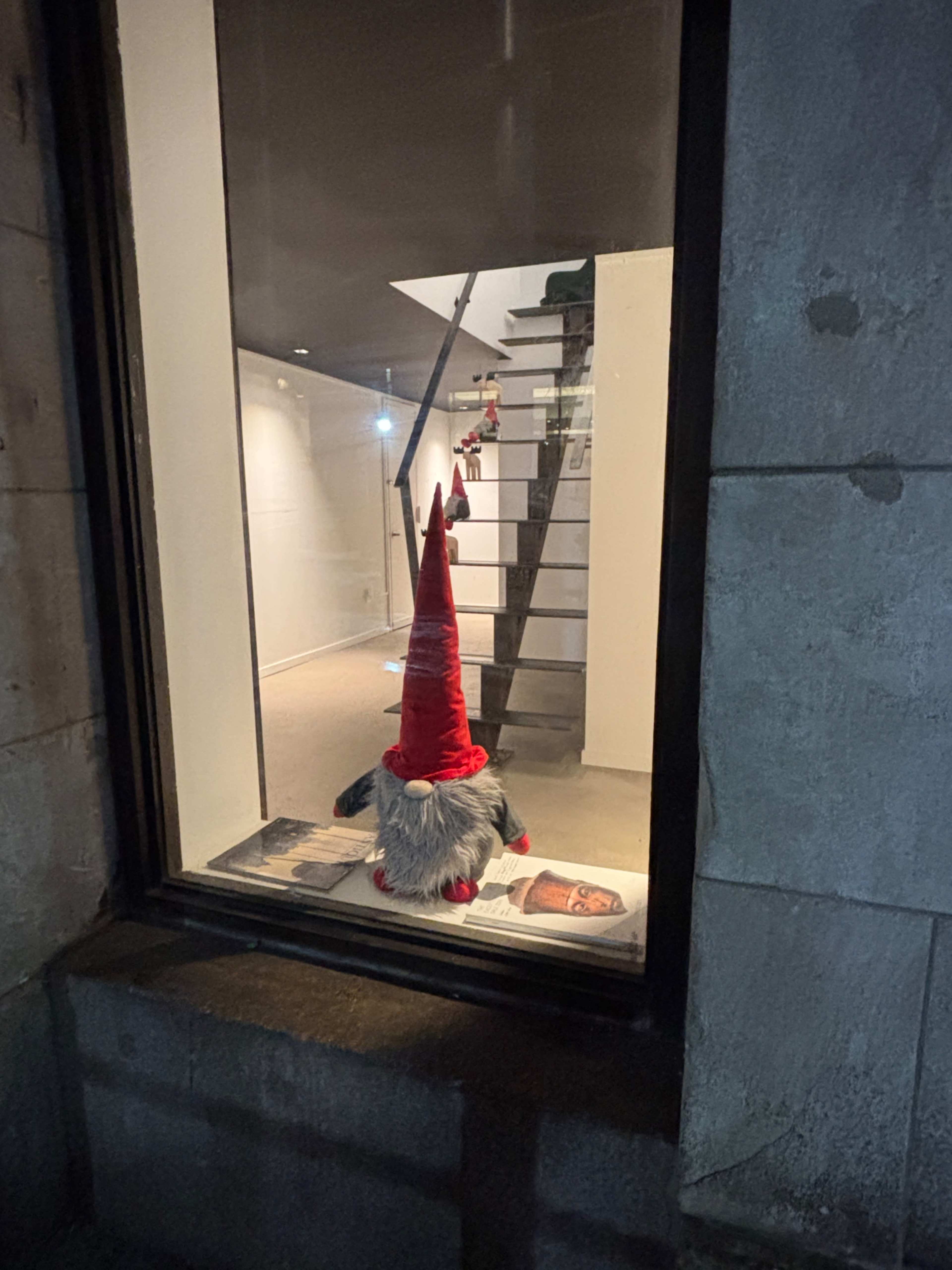 A gnome with a red hat and gray beard stands in a window, looking out at a staircase illuminated by a light above.