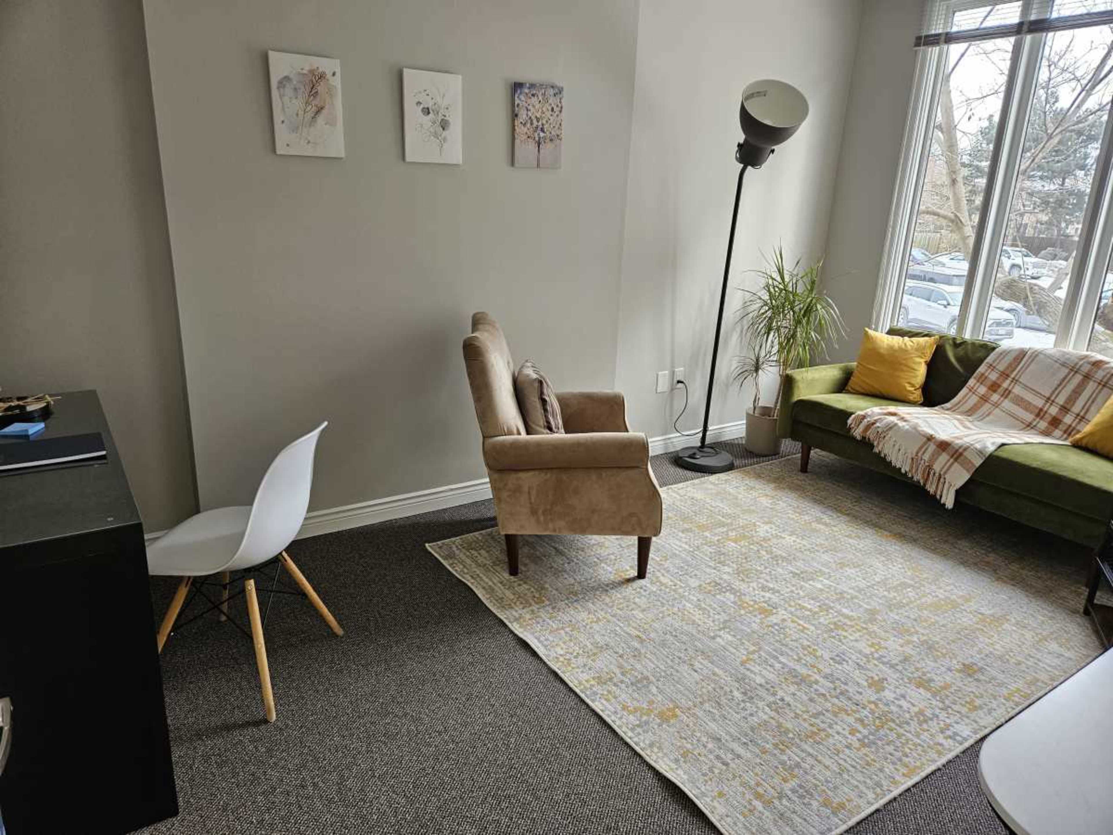 The image shows a cozy living space featuring a brown armchair, a green sofa with yellow and white pillows, a small desk, and a large area rug on carpeted flooring.