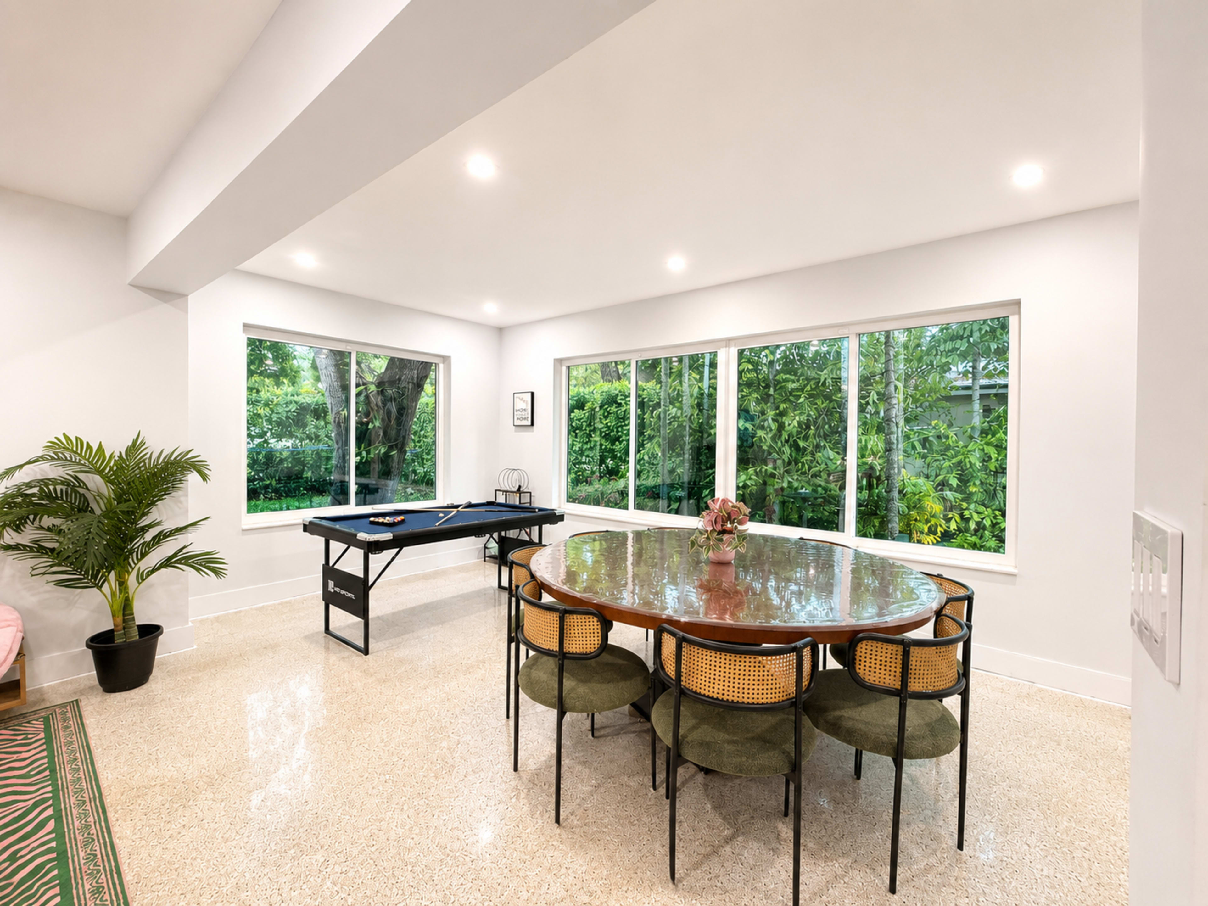 A modern dining area features a round wooden table surrounded by six chairs, with large windows offering a view of greenery and a pool table nearby.