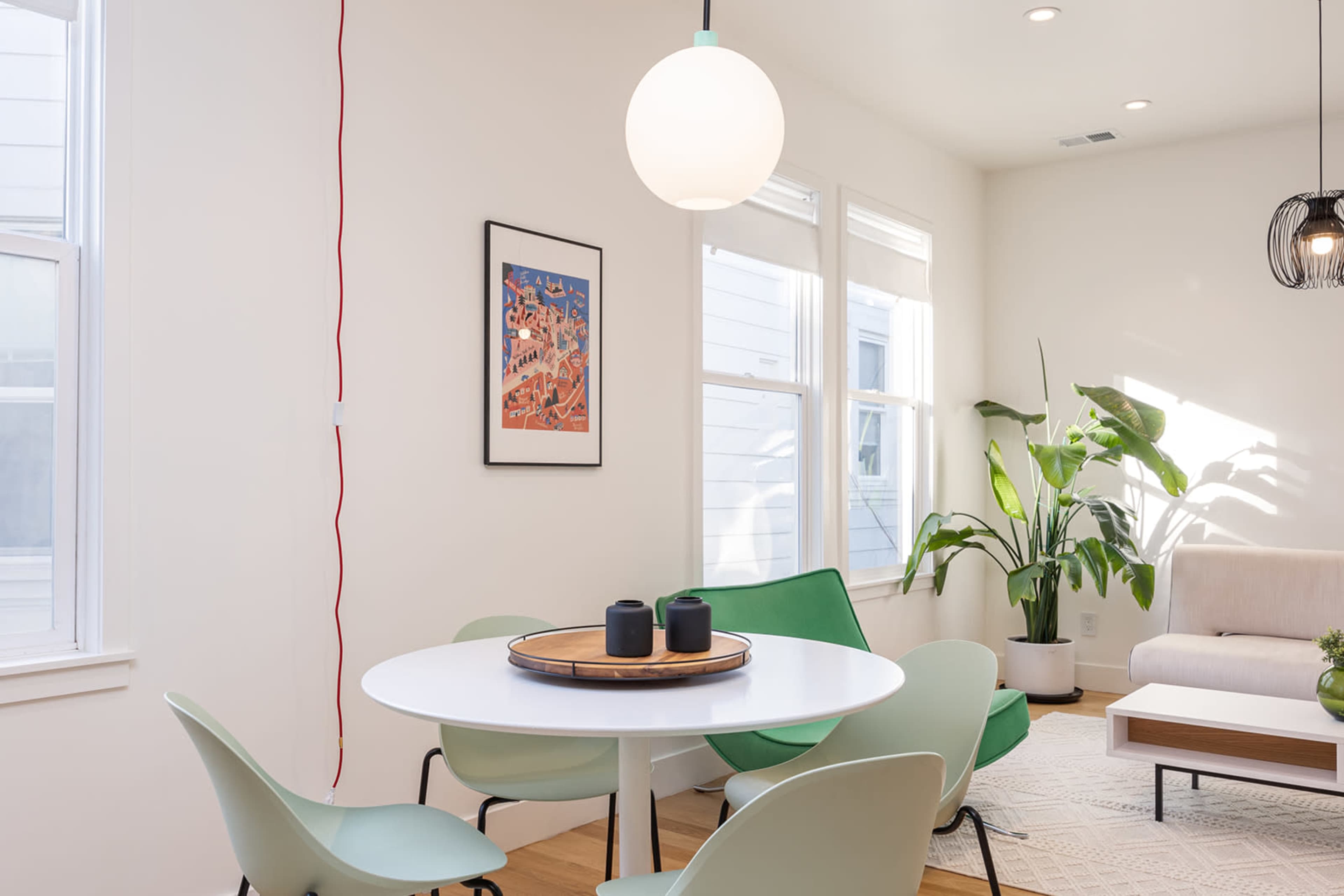 A modern living space with a round table surrounded by green chairs, a sofa, and large windows allowing natural light.