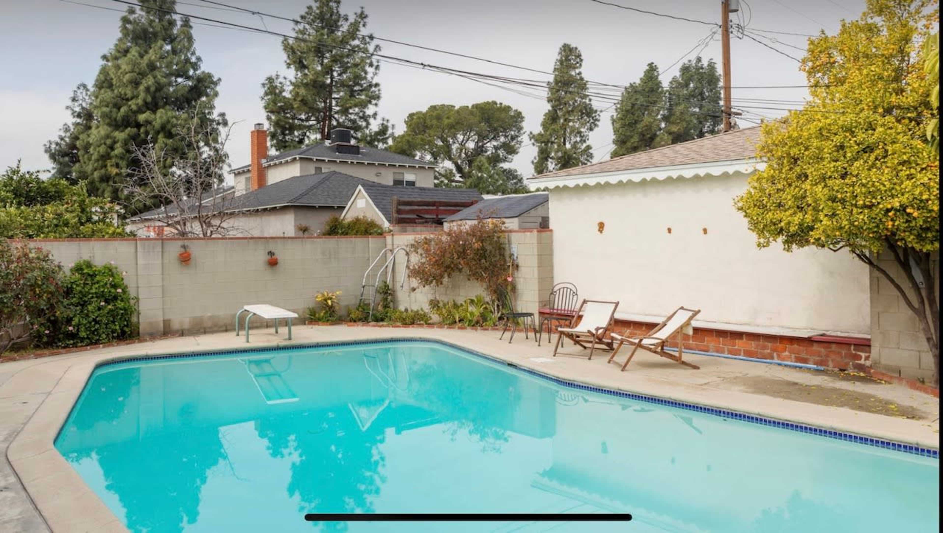 A swimming pool surrounded by concrete and greenery, with two lounge chairs positioned nearby.