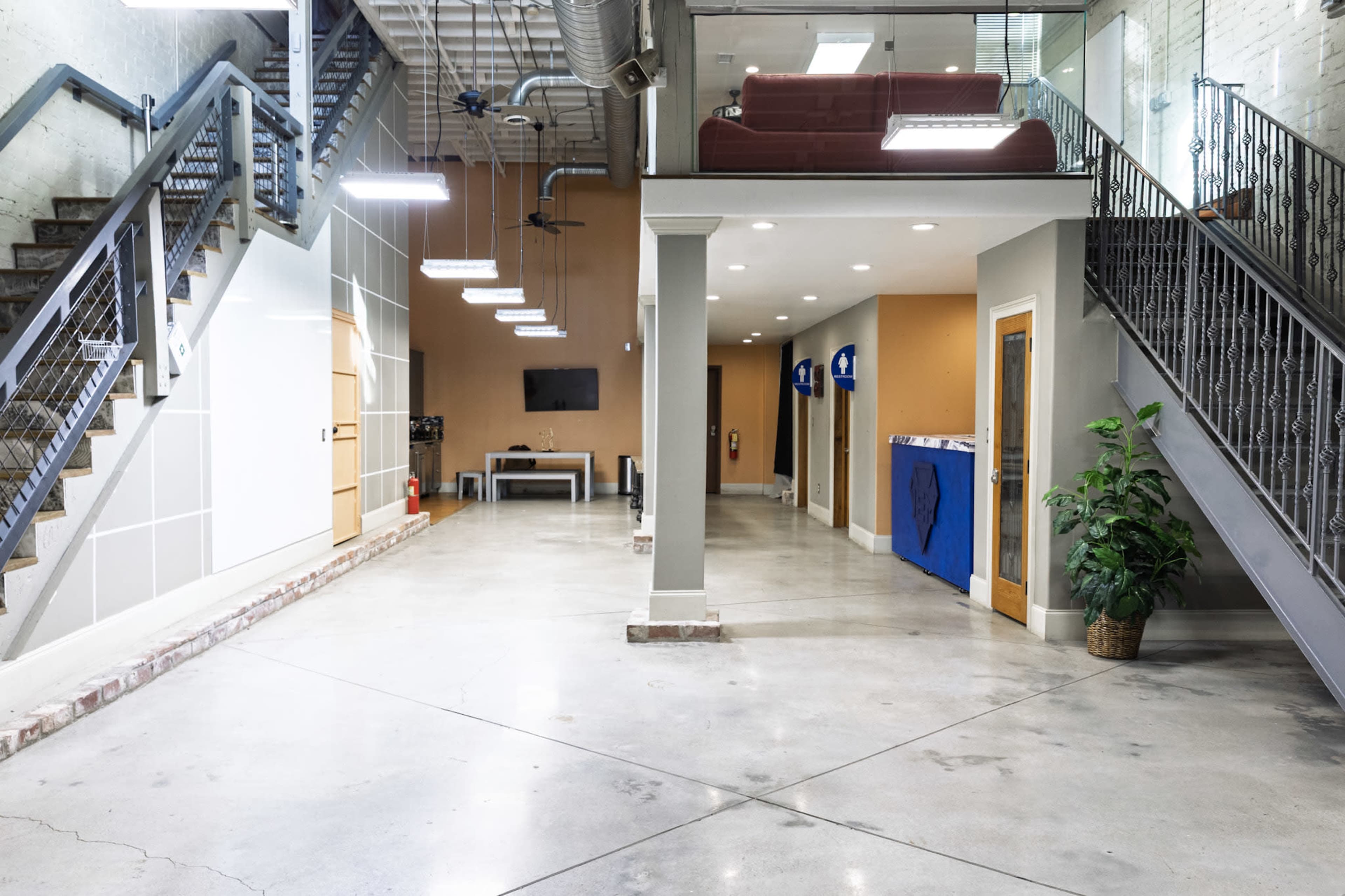 The image shows a modern indoor space with a concrete floor, featuring a staircase on the left, a seating area with a red couch on an upper level, and a reception area with office doors on the right.