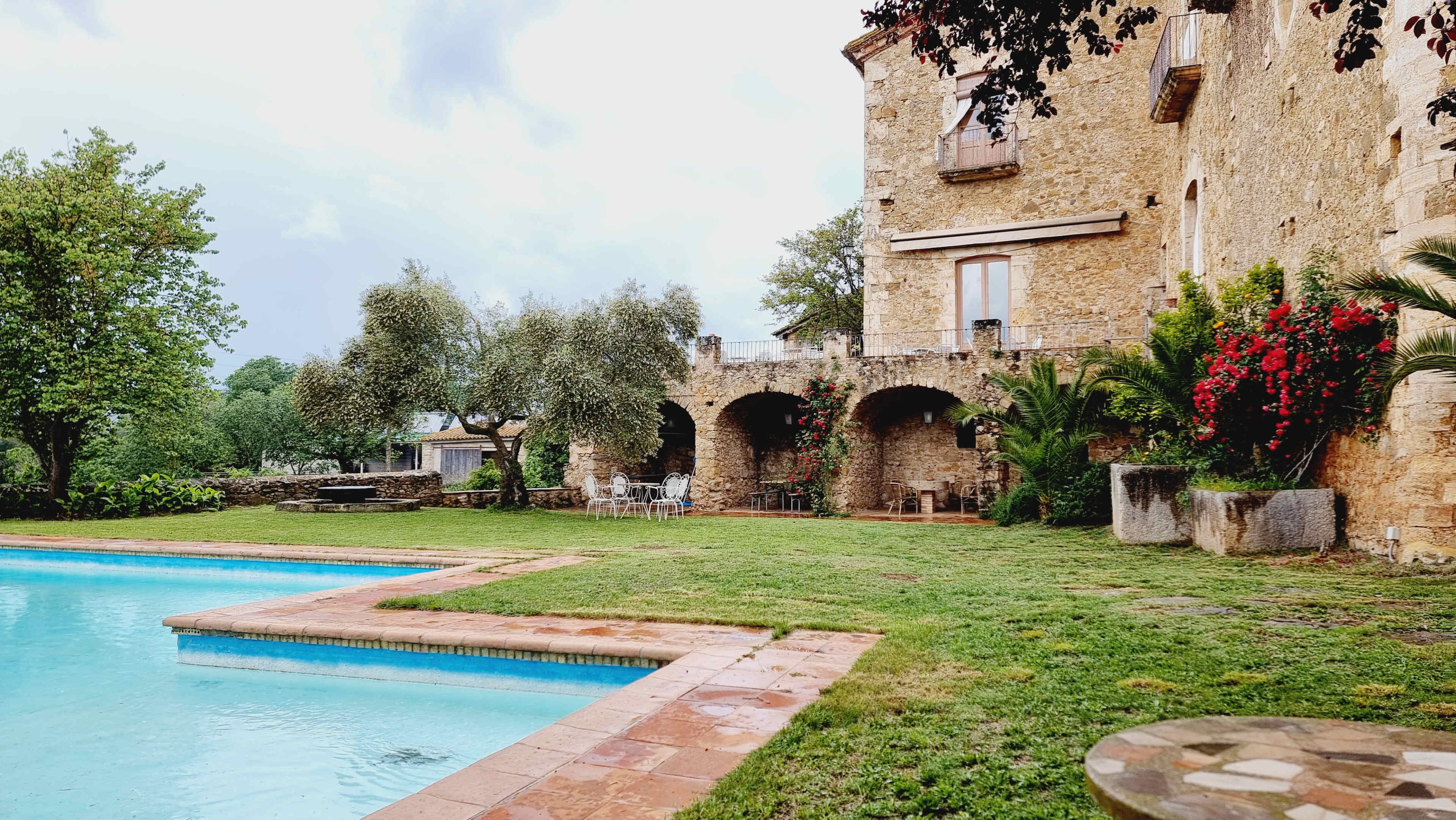 A stone building with balconies overlooks a tidy lawn and a rectangular swimming pool surrounded by greenery.