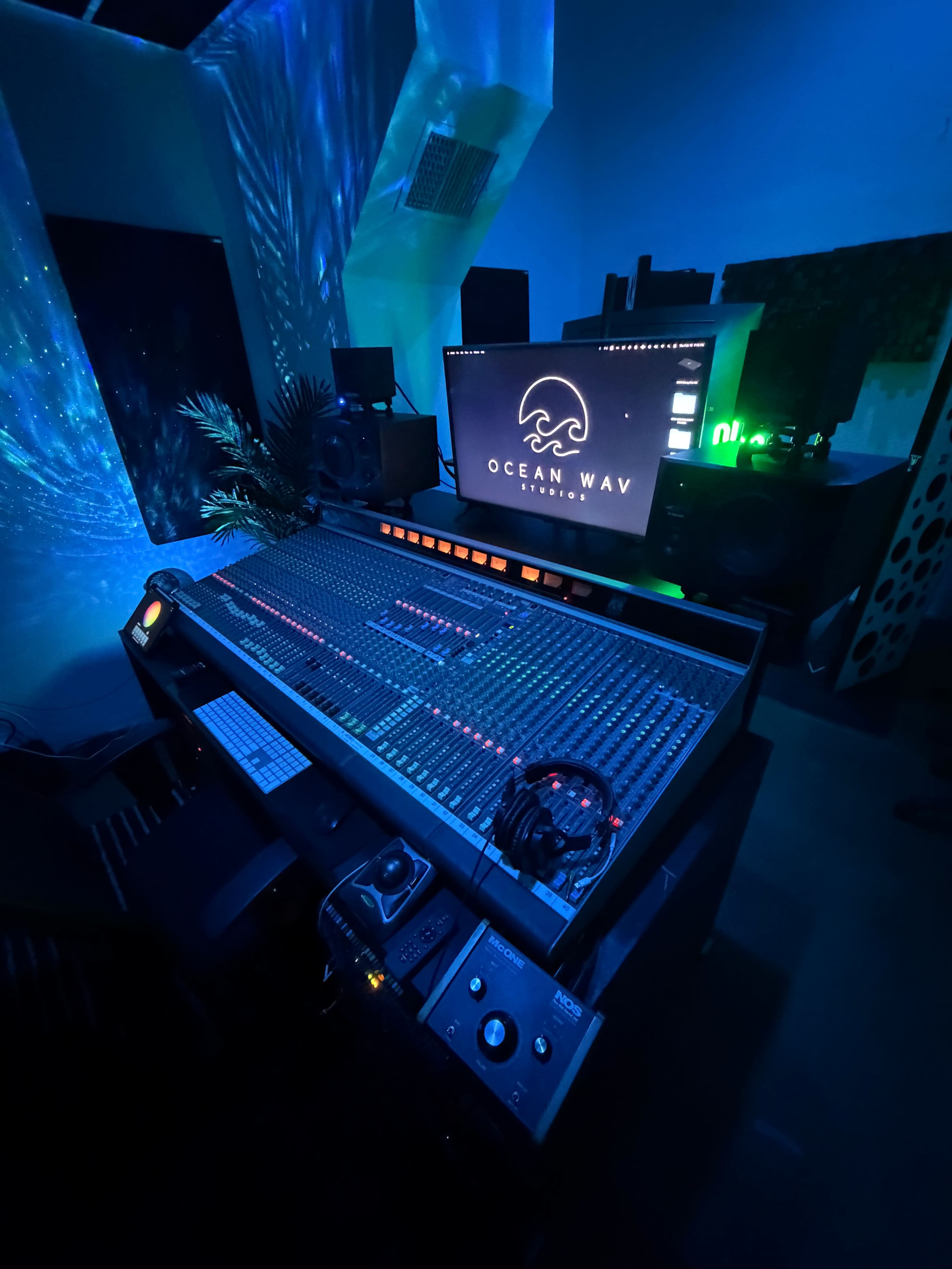 The image shows a music production studio featuring a large mixing console, monitors, and ambient lighting.