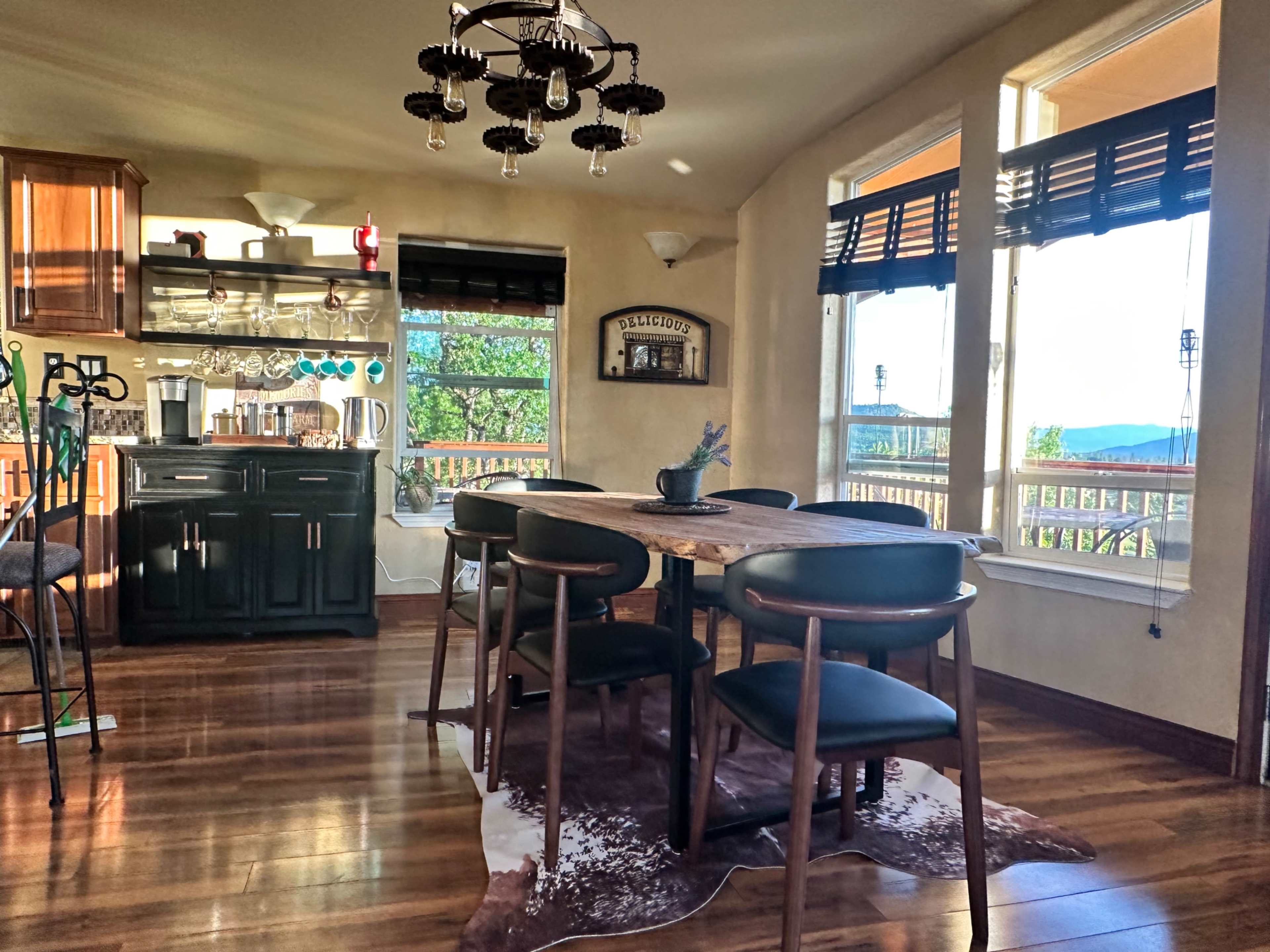 A dining area features a wooden table surrounded by black chairs, with a kitchen area visible in the background and large windows letting in natural light.