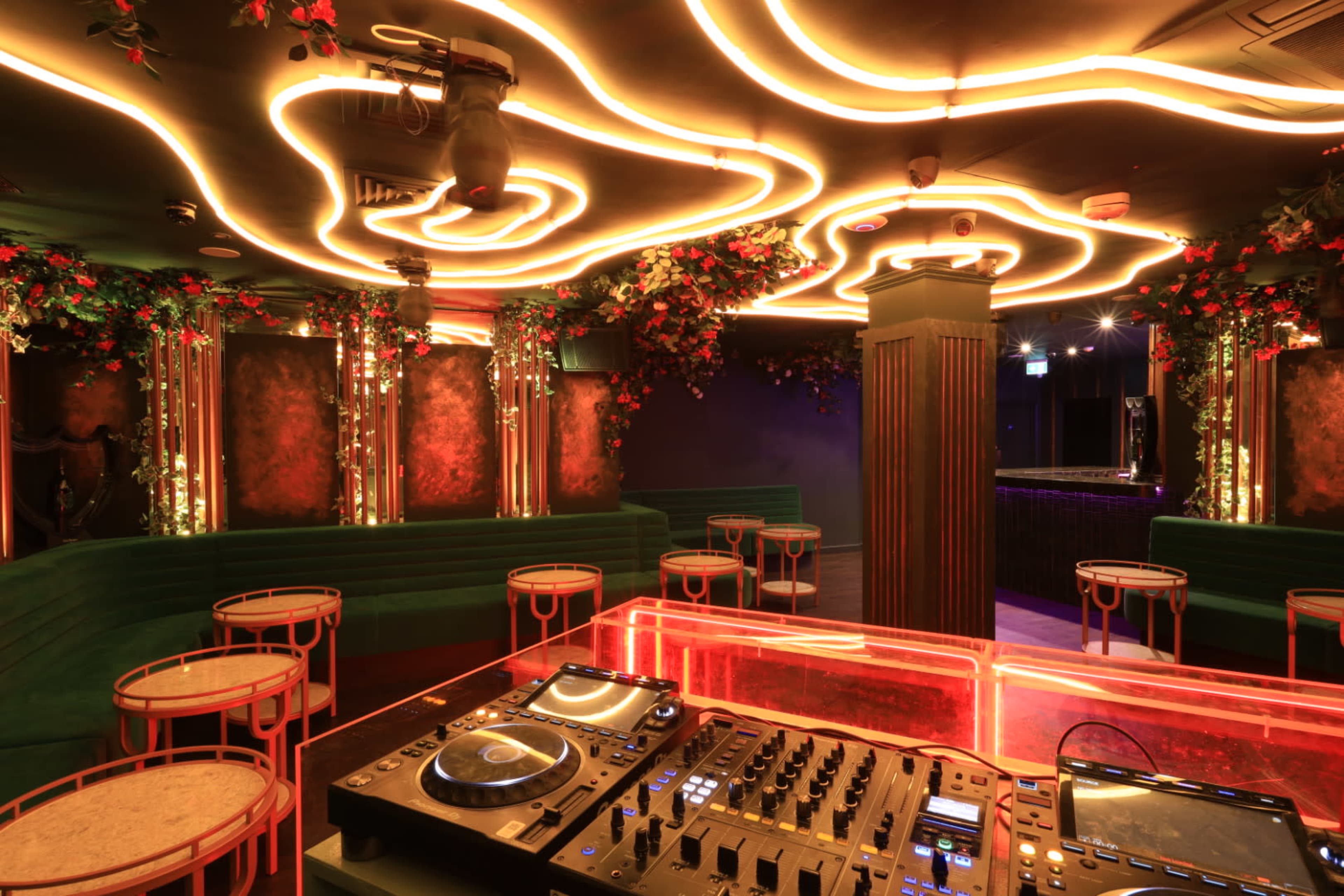 The image shows a dimly lit lounge with glowing orange ceiling designs and green seating, featuring a DJ equipment setup in the foreground.