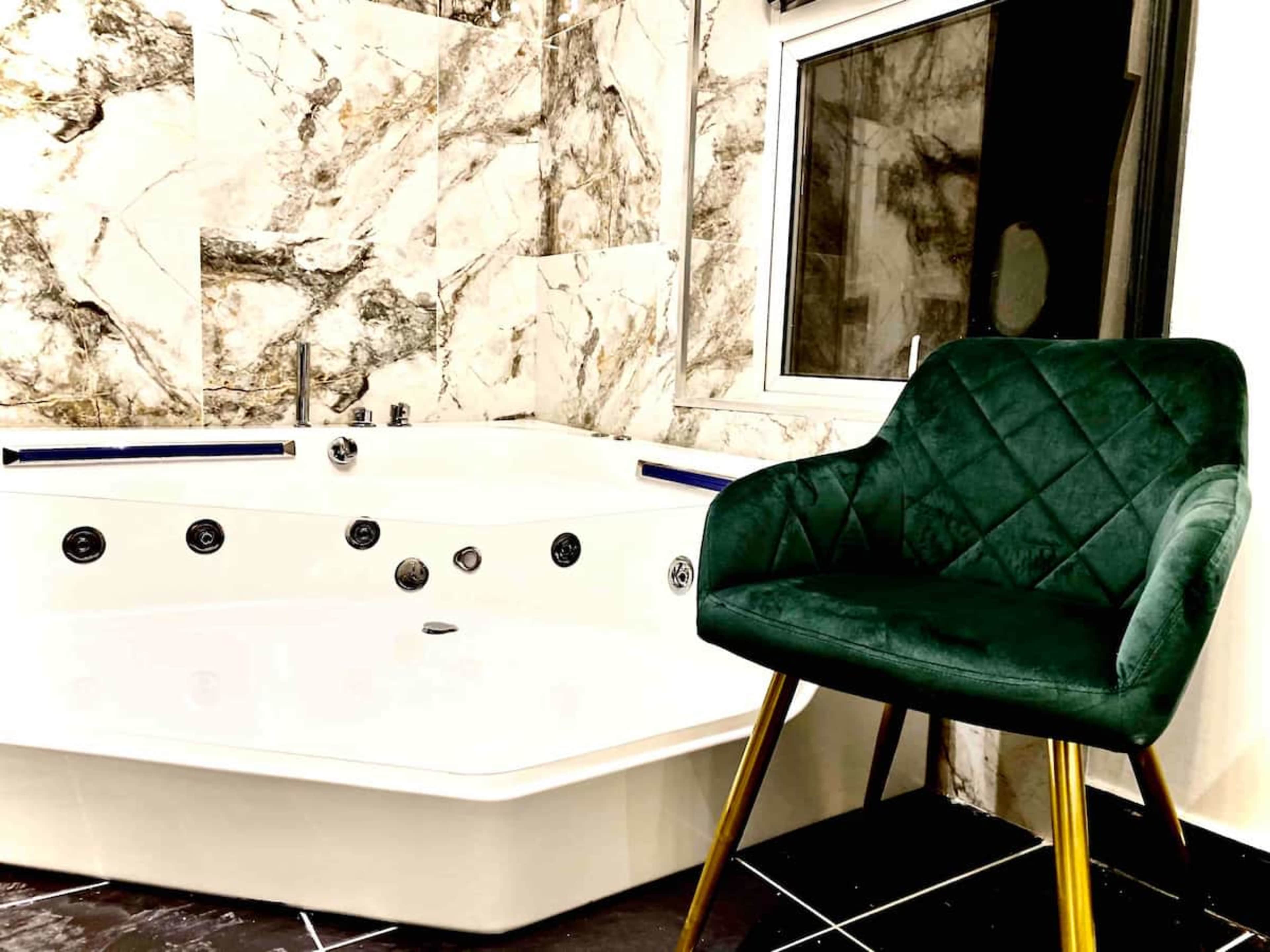 A modern bathroom features a white jacuzzi bathtub next to a green velvet chair with gold legs.