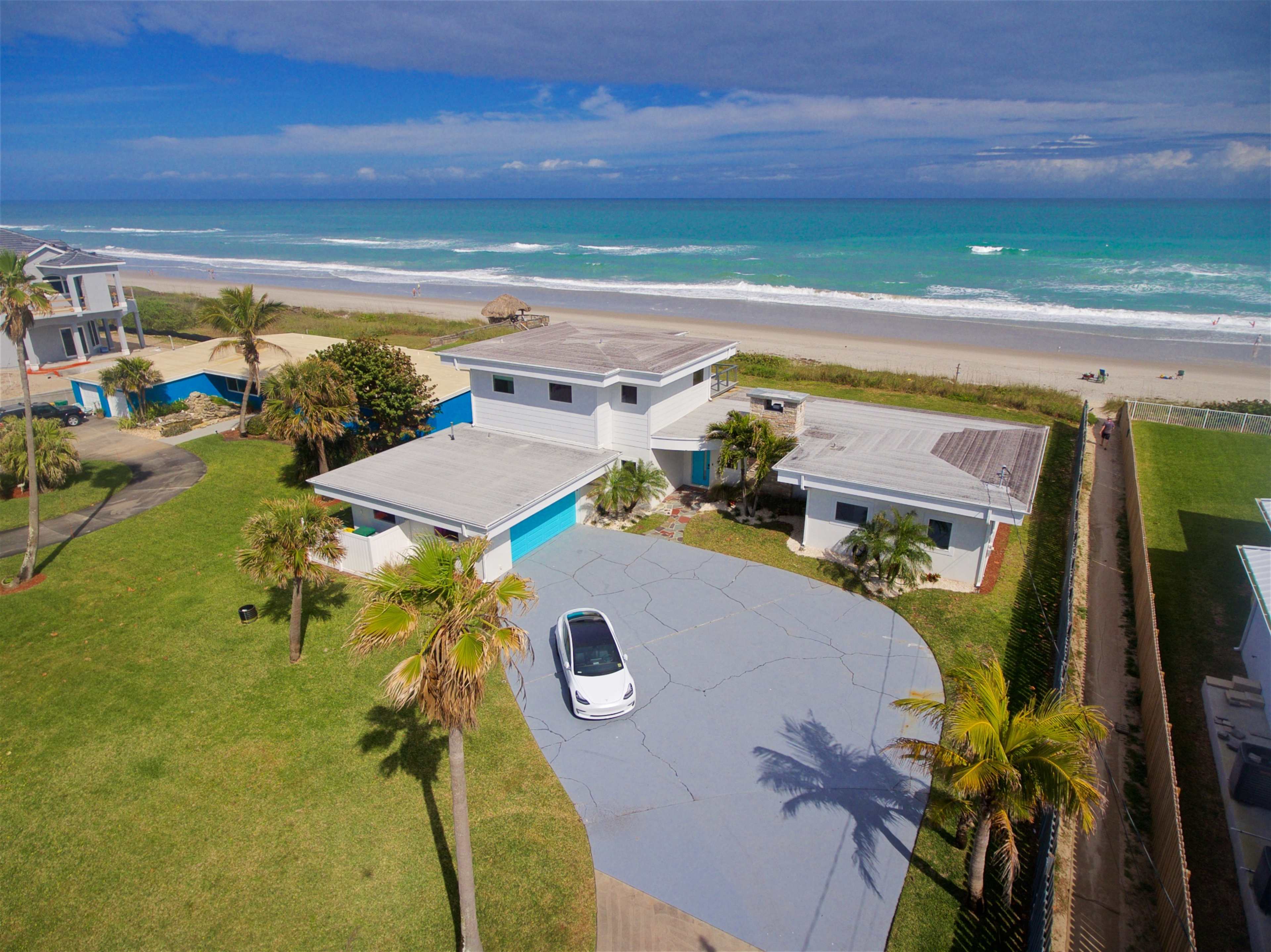A beachfront property with two houses, a concrete driveway, palm trees, and the ocean in the background.