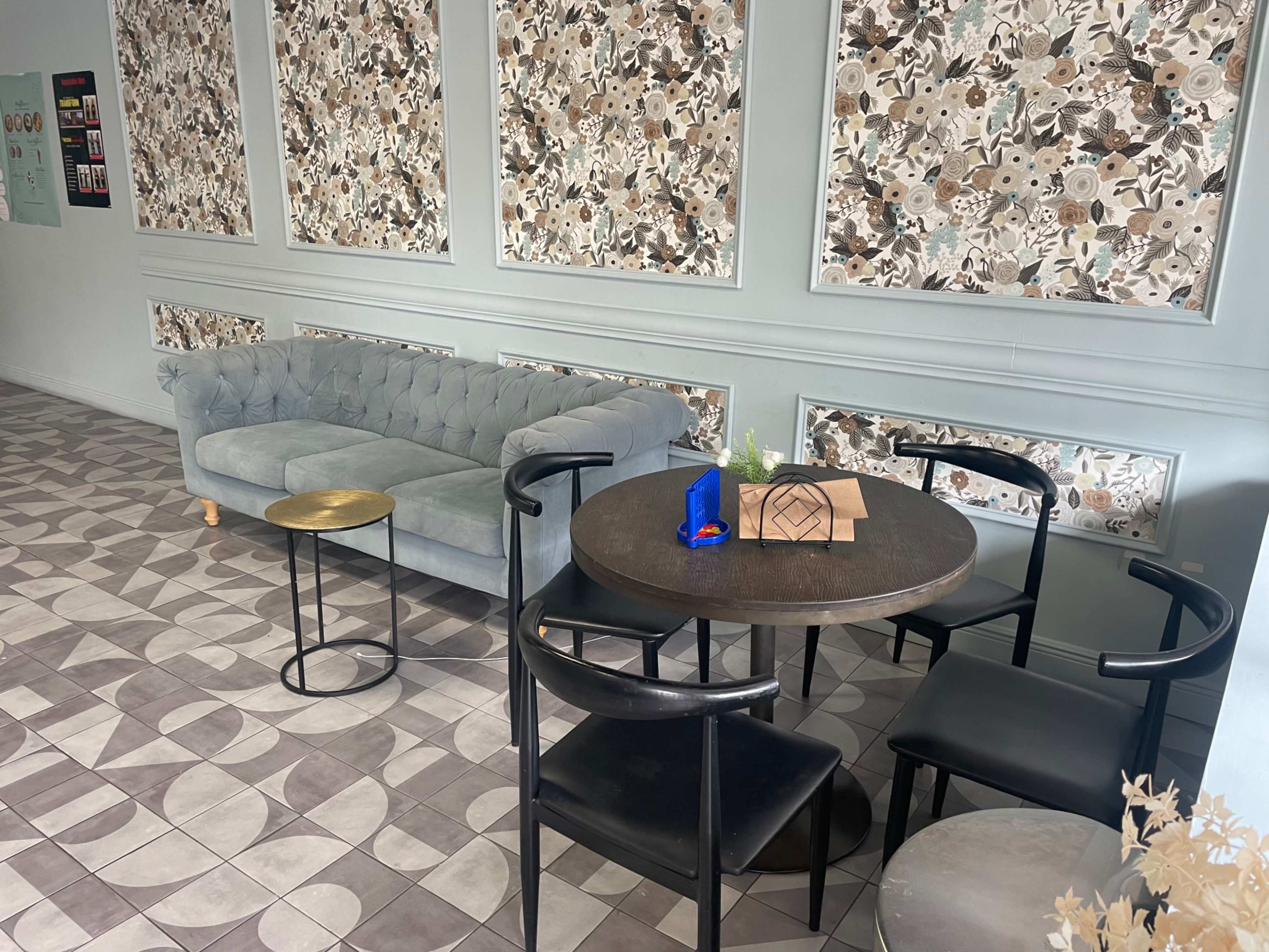 The image shows a cozy café interior with a tufted light blue couch, a round black table surrounded by black chairs, and decorative wall panels featuring floral patterns.