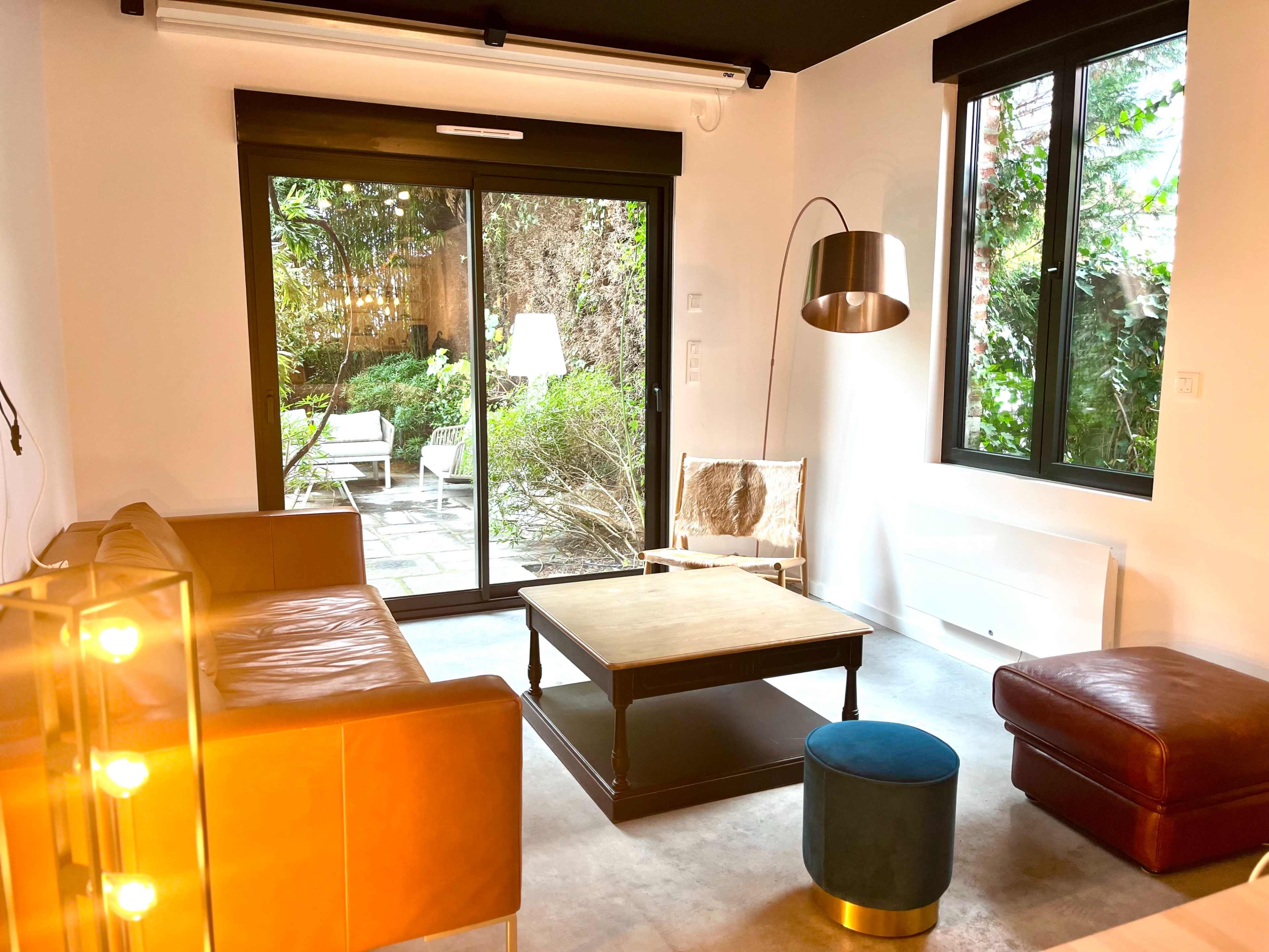 The image shows a cozy living room with leather sofas, a wooden coffee table, and large windows overlooking a garden.