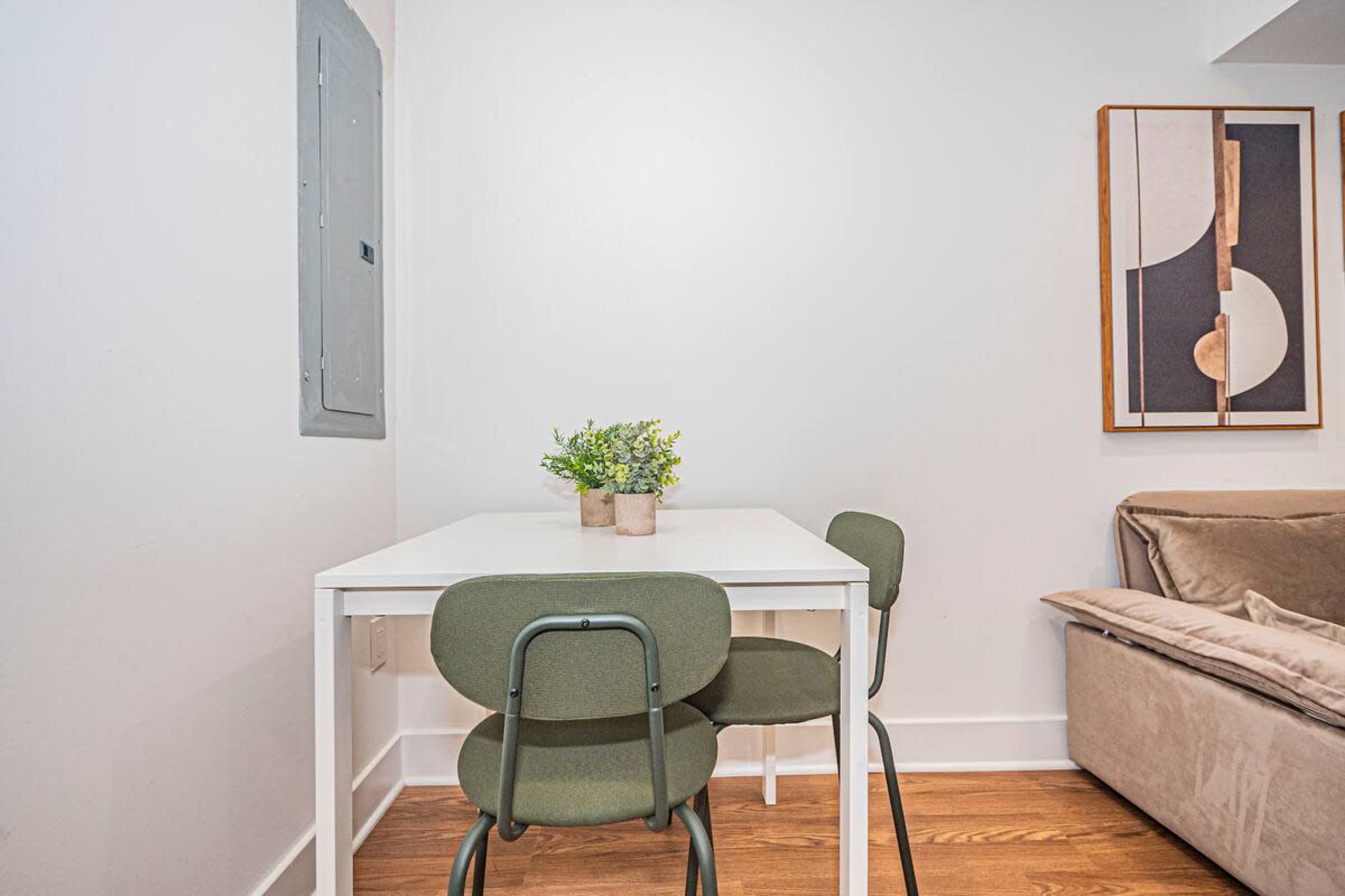 A small dining table with two green chairs and a potted plant is positioned against a white wall, near a couch and a wall-mounted artwork.