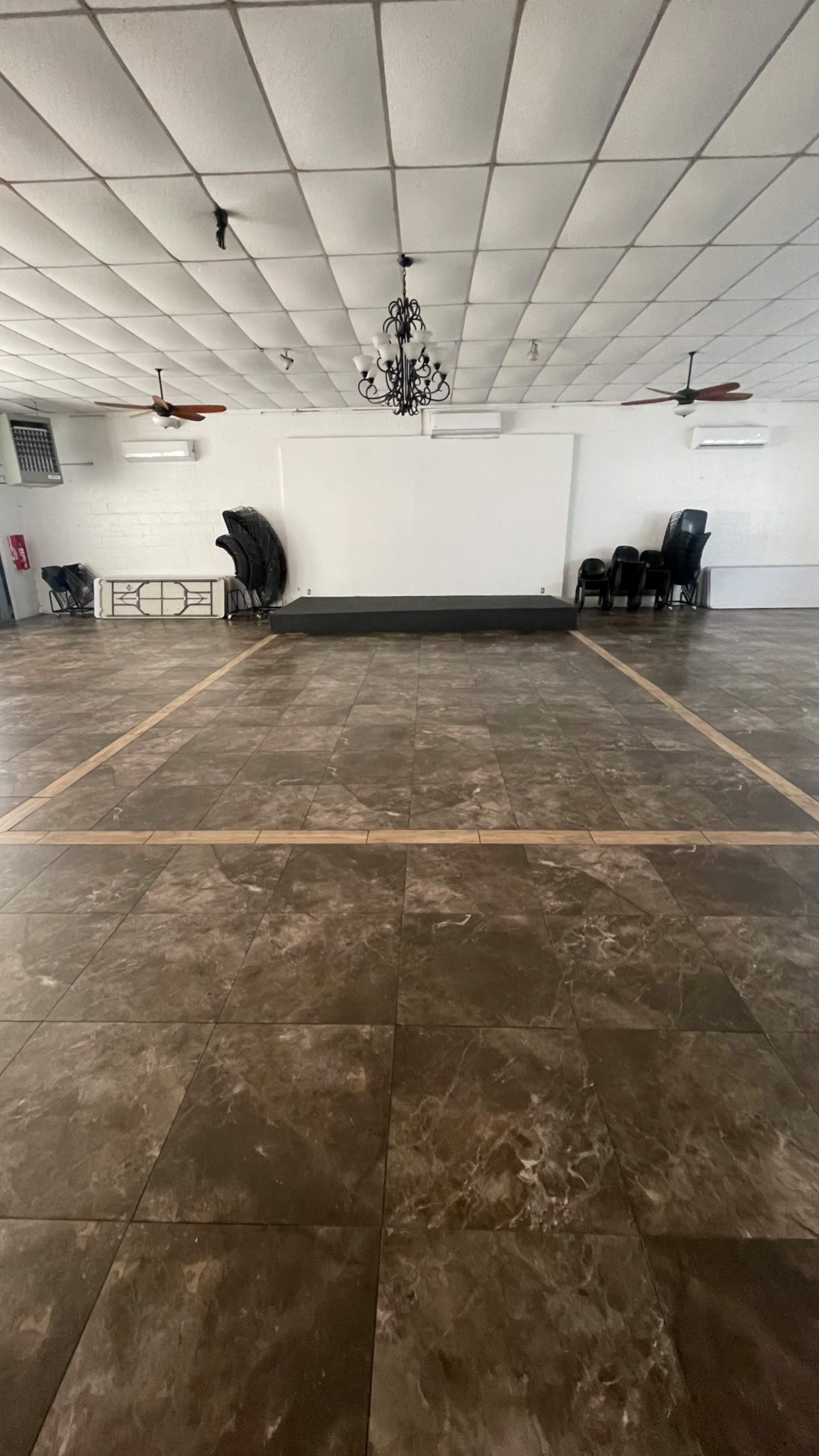 The image shows a spacious hall with a tiled floor, a central stage area, and rows of black chairs arranged along the perimeter.