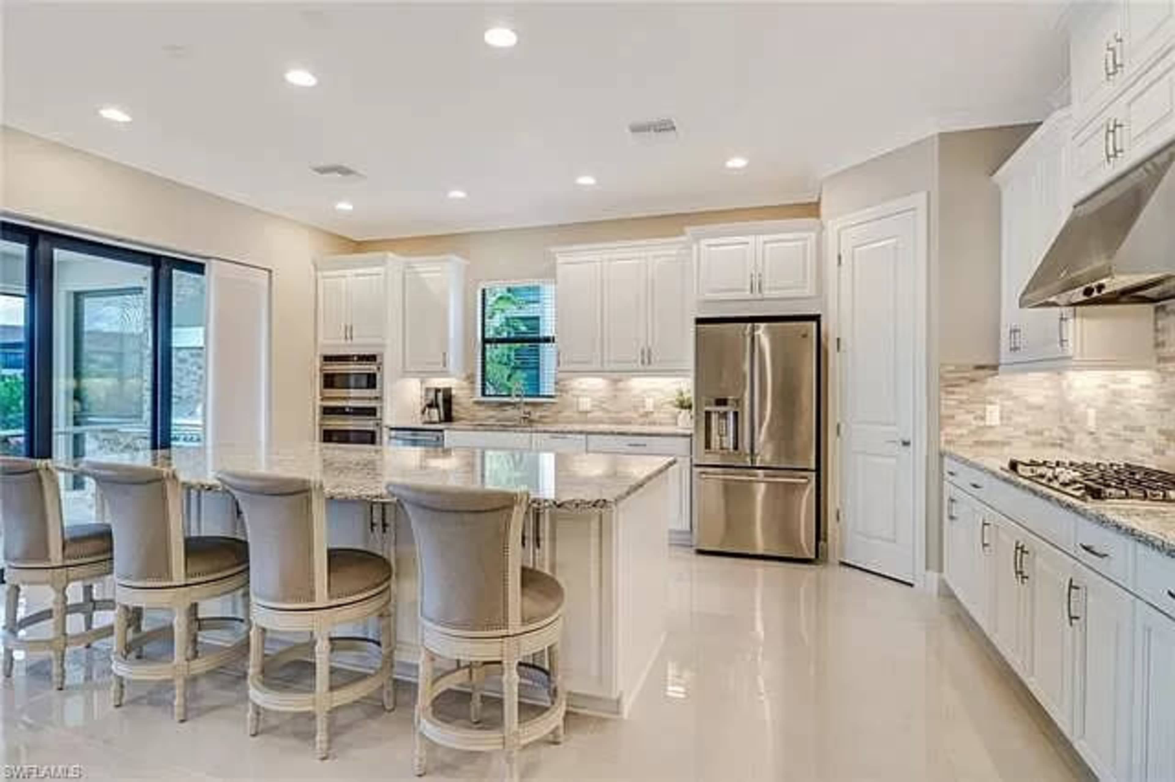 A modern kitchen features white cabinetry, a large island with bar seating, stainless steel appliances, and natural light from large windows.