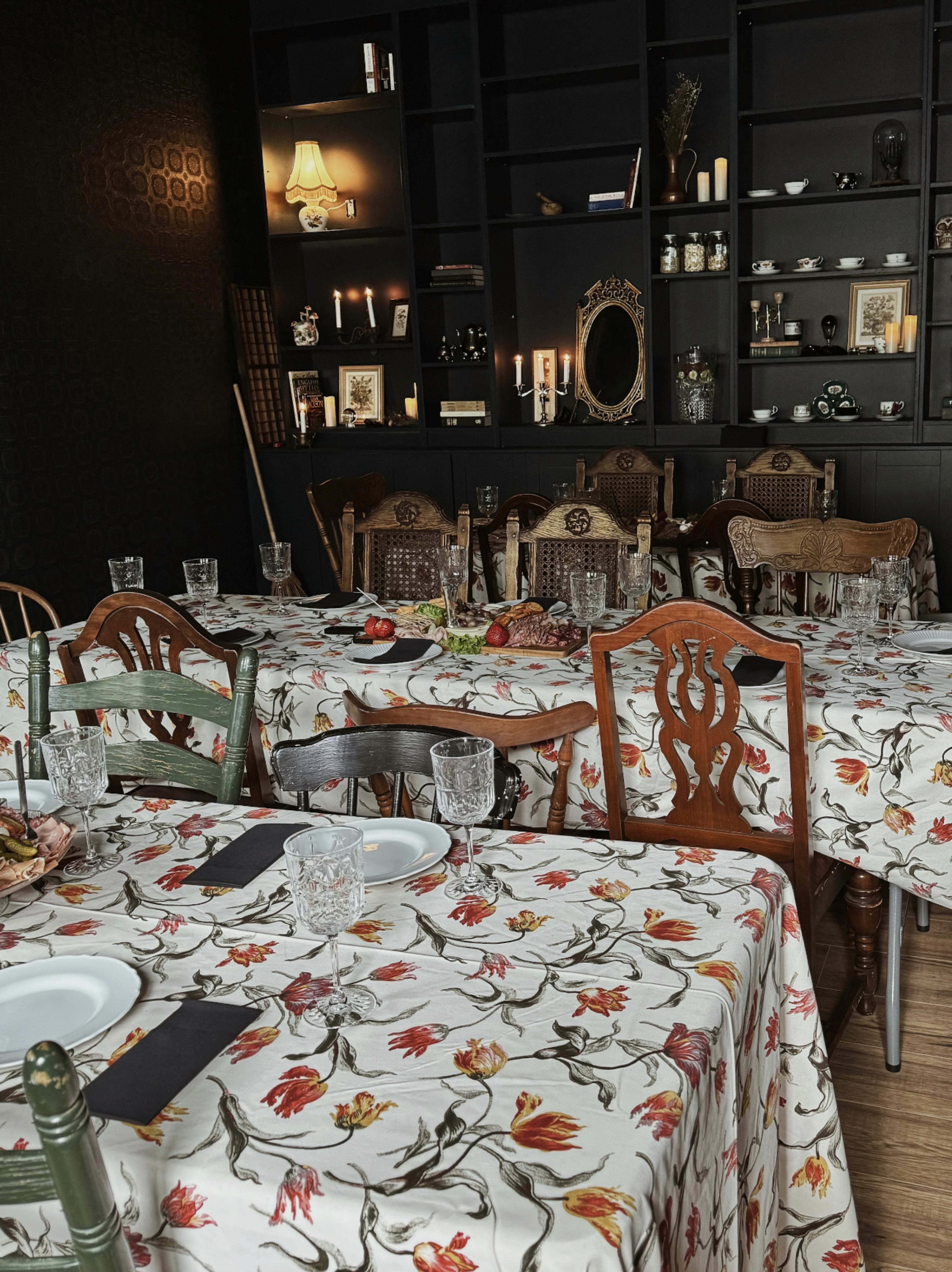 The image depicts a restaurant interior with several tables covered in floral-patterned tablecloths, arranged neatly in a dimly lit space featuring shelves filled with decor and vintage items.