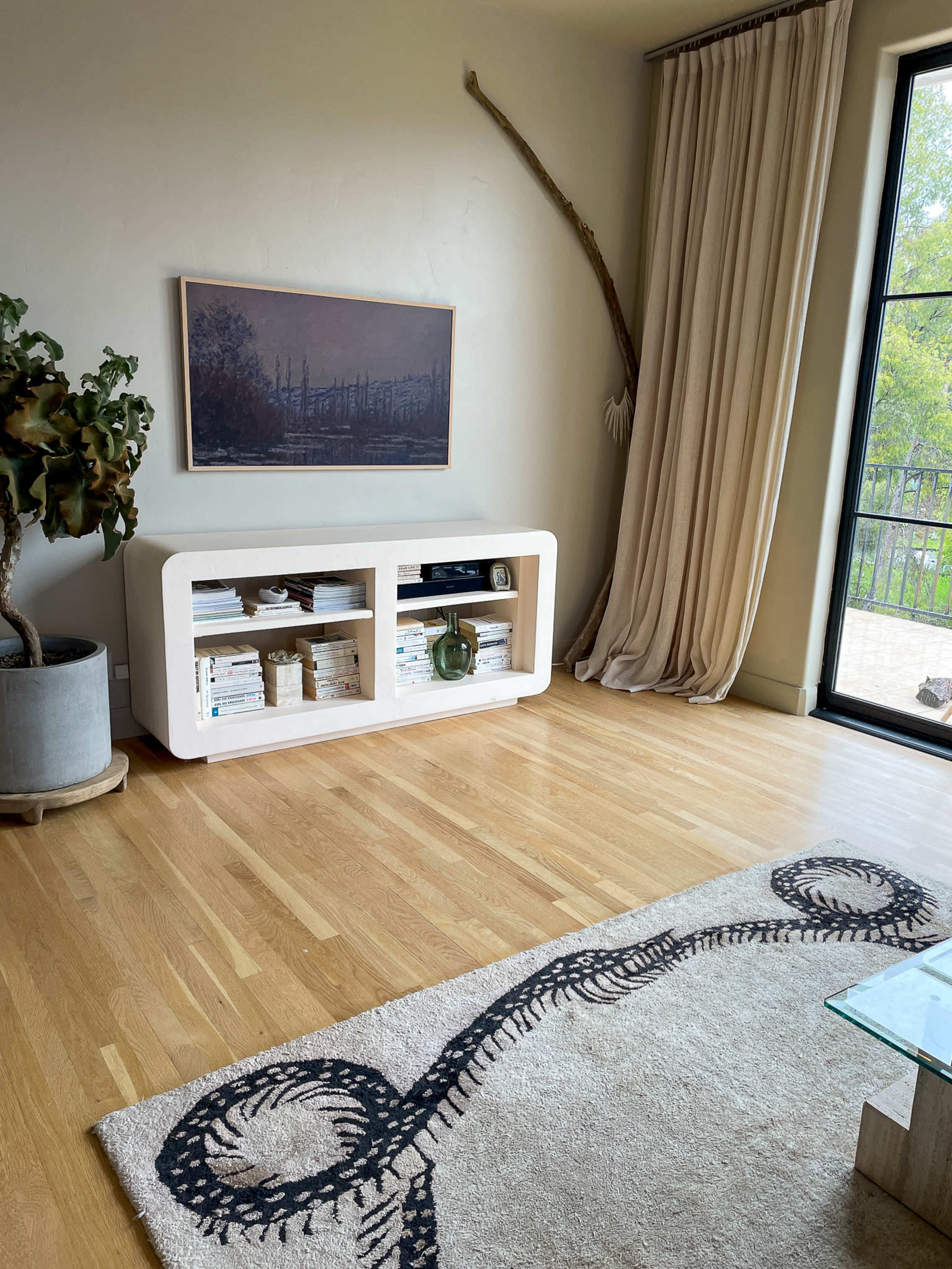 The image shows a living room corner with a light-colored console table displaying books and decor, a potted plant, a large curtain, and a window overlooking an outdoor area.