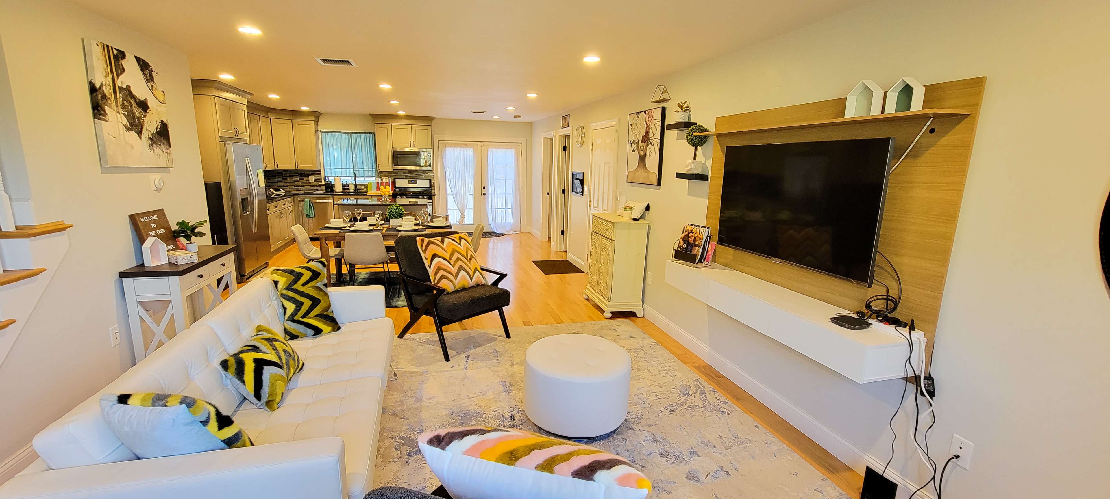 A modern living room with a white sectional sofa, decorative cushions, a wall-mounted television, and an open layout leading to a kitchen area.