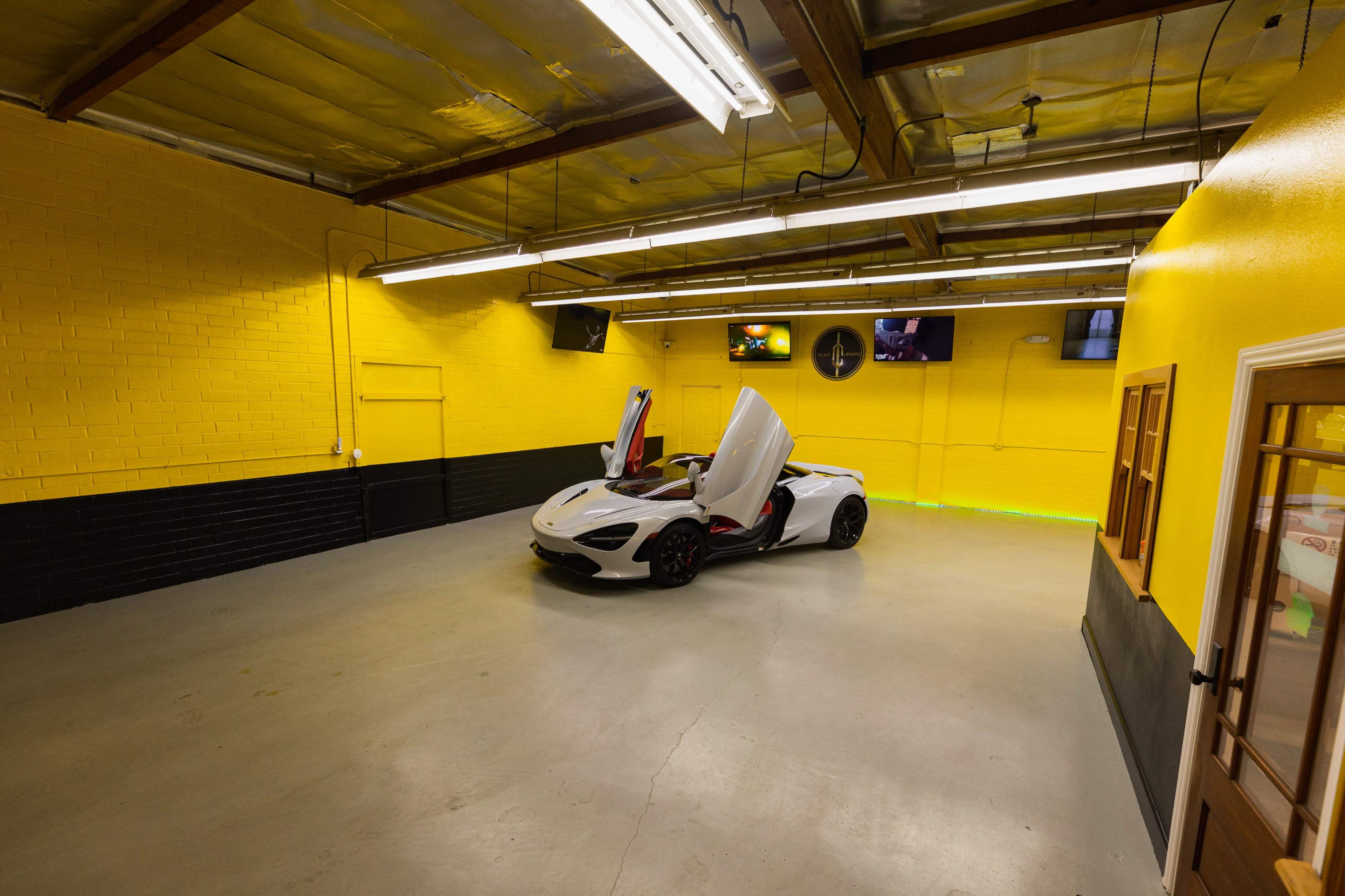 A white McLaren 720S with its doors raised is parked in a spacious, brightly lit garage with yellow walls and black accents.