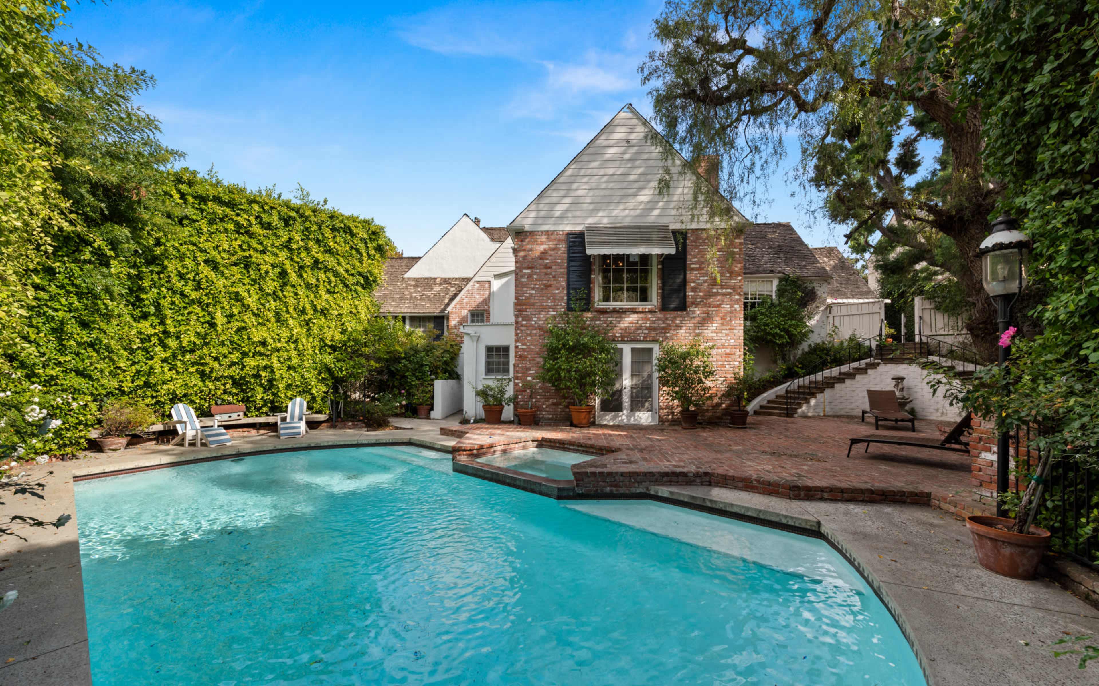 A backyard with a swimming pool, surrounded by greenery and a brick patio adjacent to a two-story house.