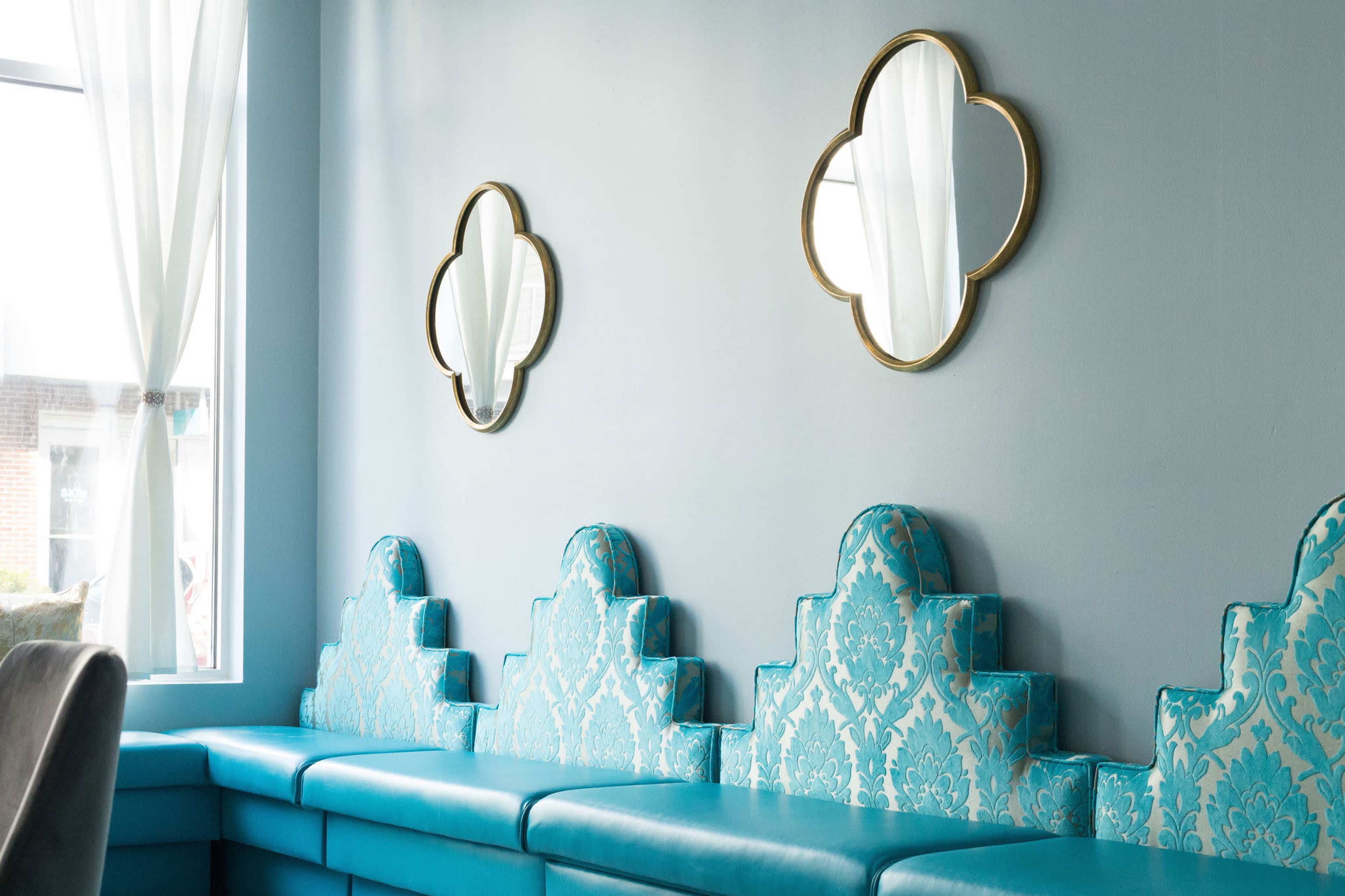 A light blue wall with two decorative mirrors above a turquoise cushioned bench that has a scalloped design.