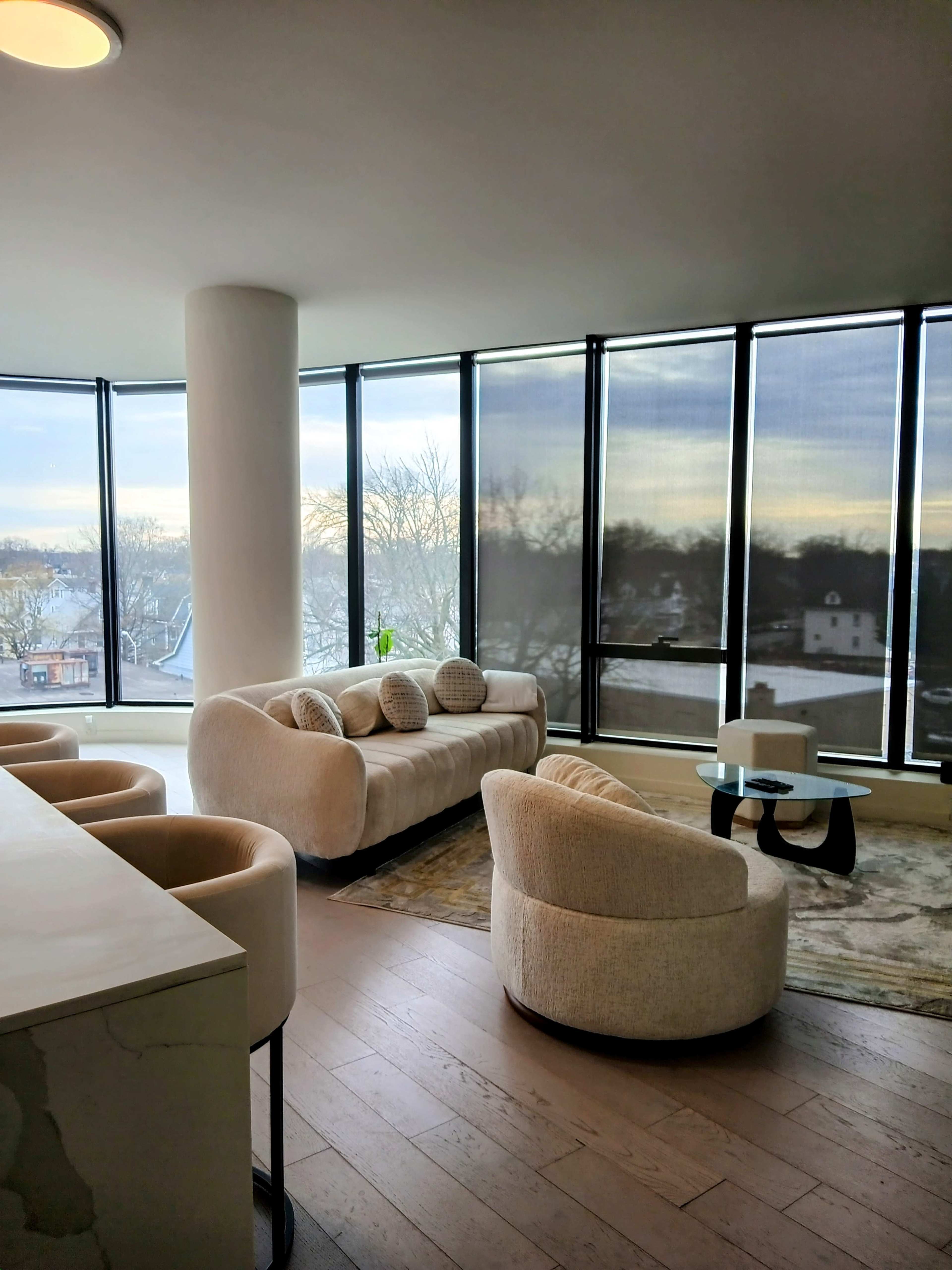 The image shows a modern, open-concept living space with large windows, featuring a beige sofa, two round chairs, and a circular glass coffee table.
