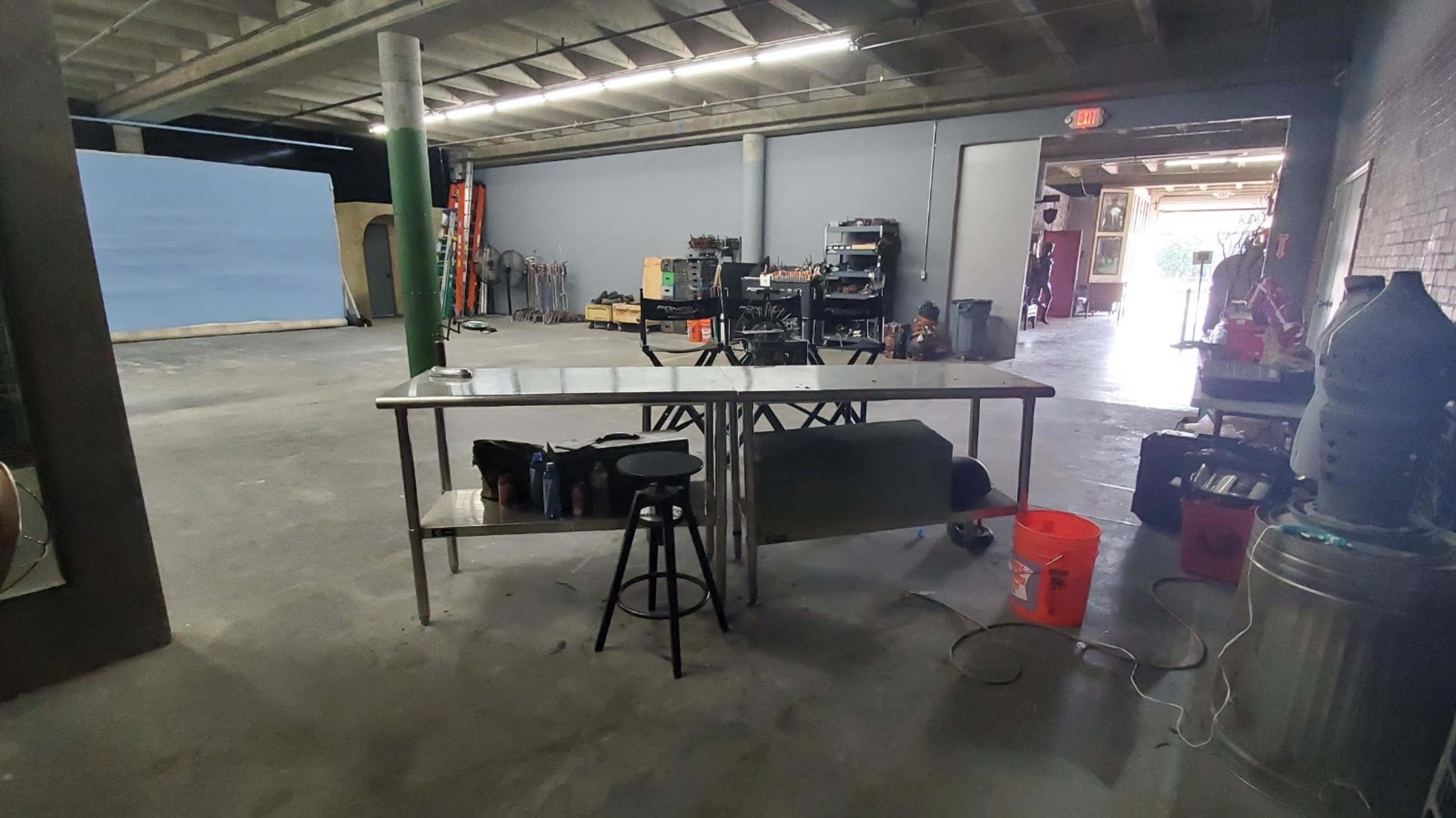 An industrial workspace featuring a metal table, a stool, a large orange bucket, and various tools and equipment along the walls.
