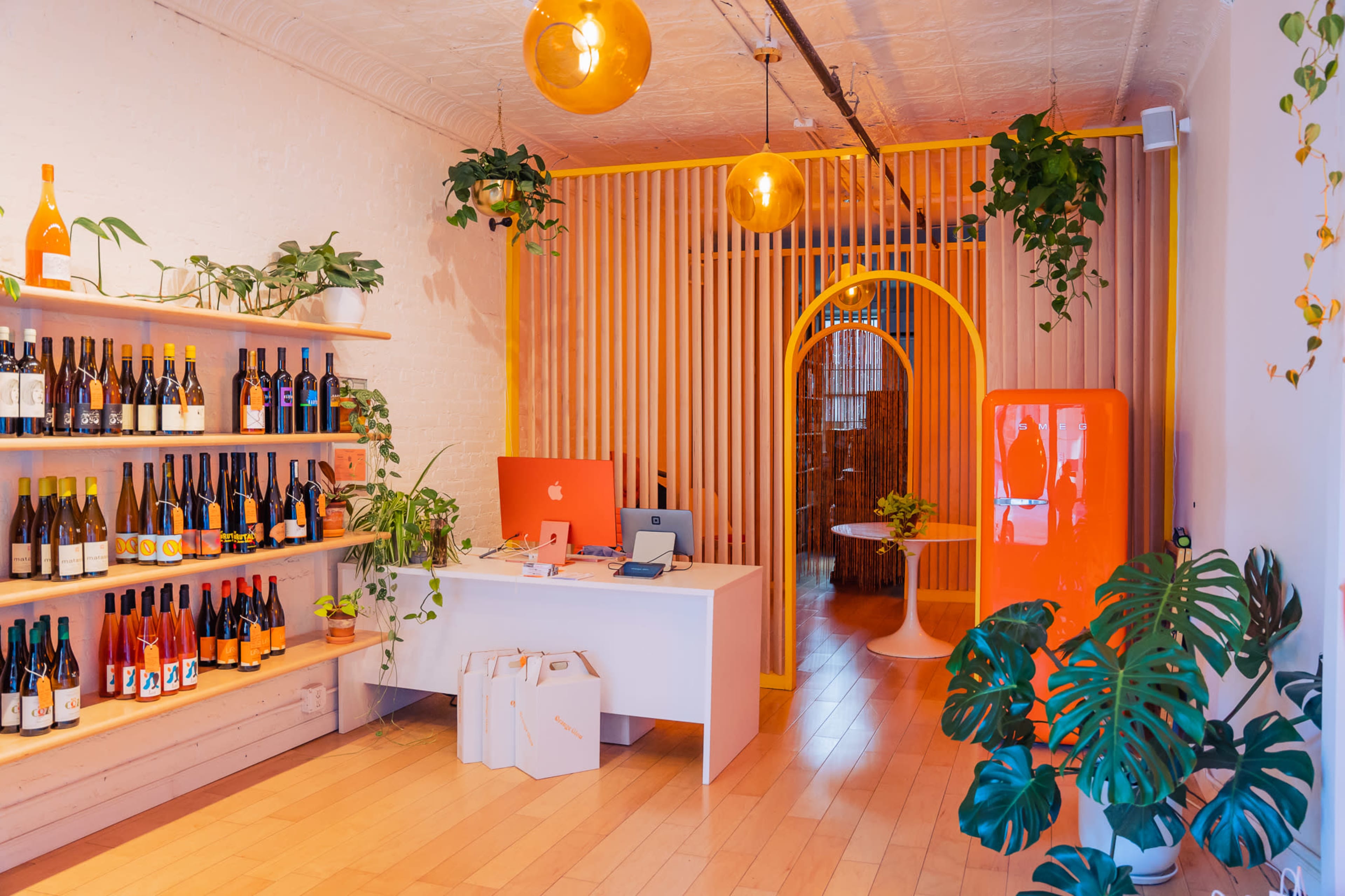 A brightly decorated retail space with shelves of wine bottles, a white reception desk with an iMac, and an archway leading to a seating area, surrounded by various plants and vibrant orange accents.