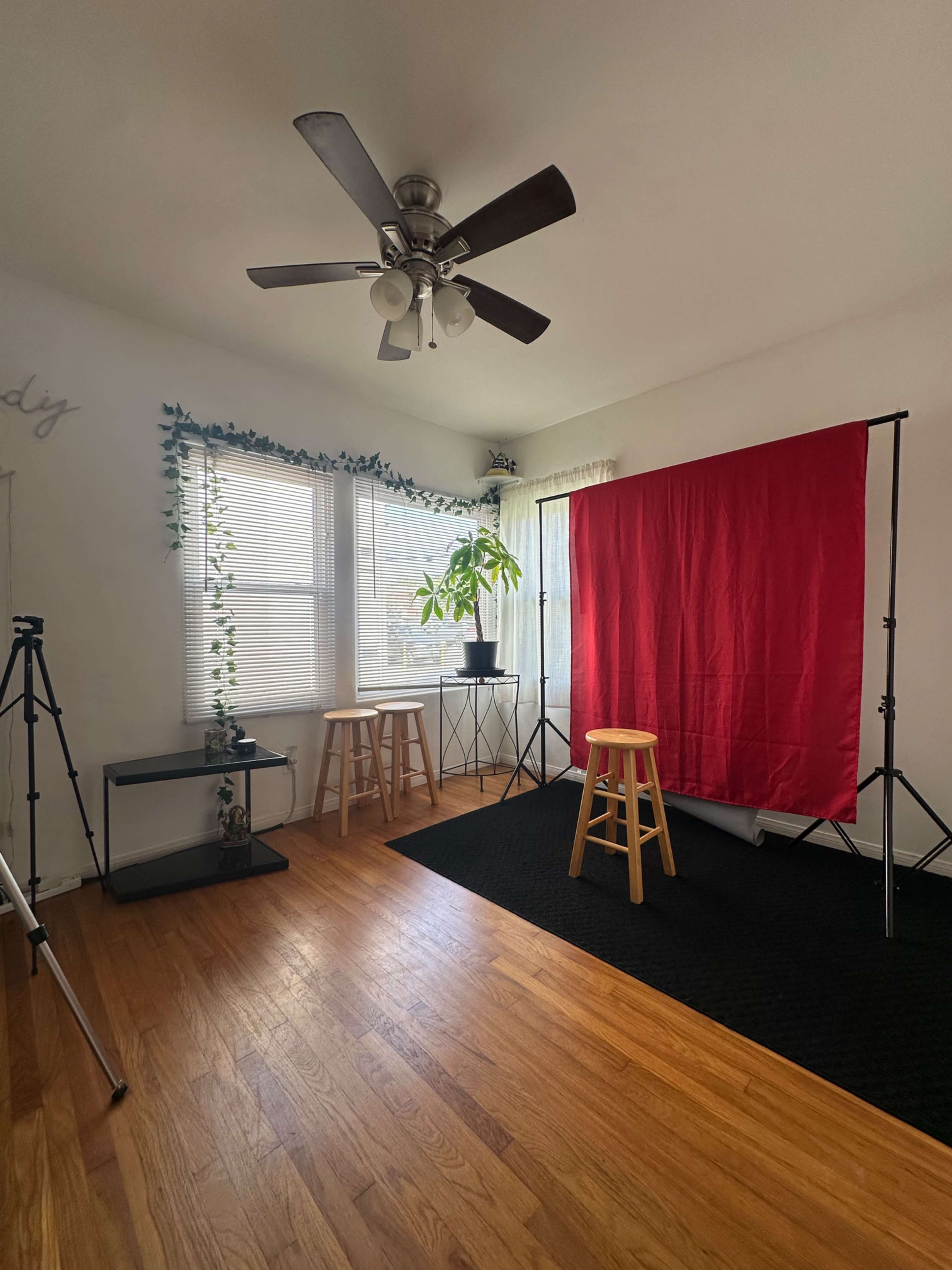 The image shows a well-lit room with a red backdrop set up for photography, two wooden stools, and various equipment arranged along the walls.