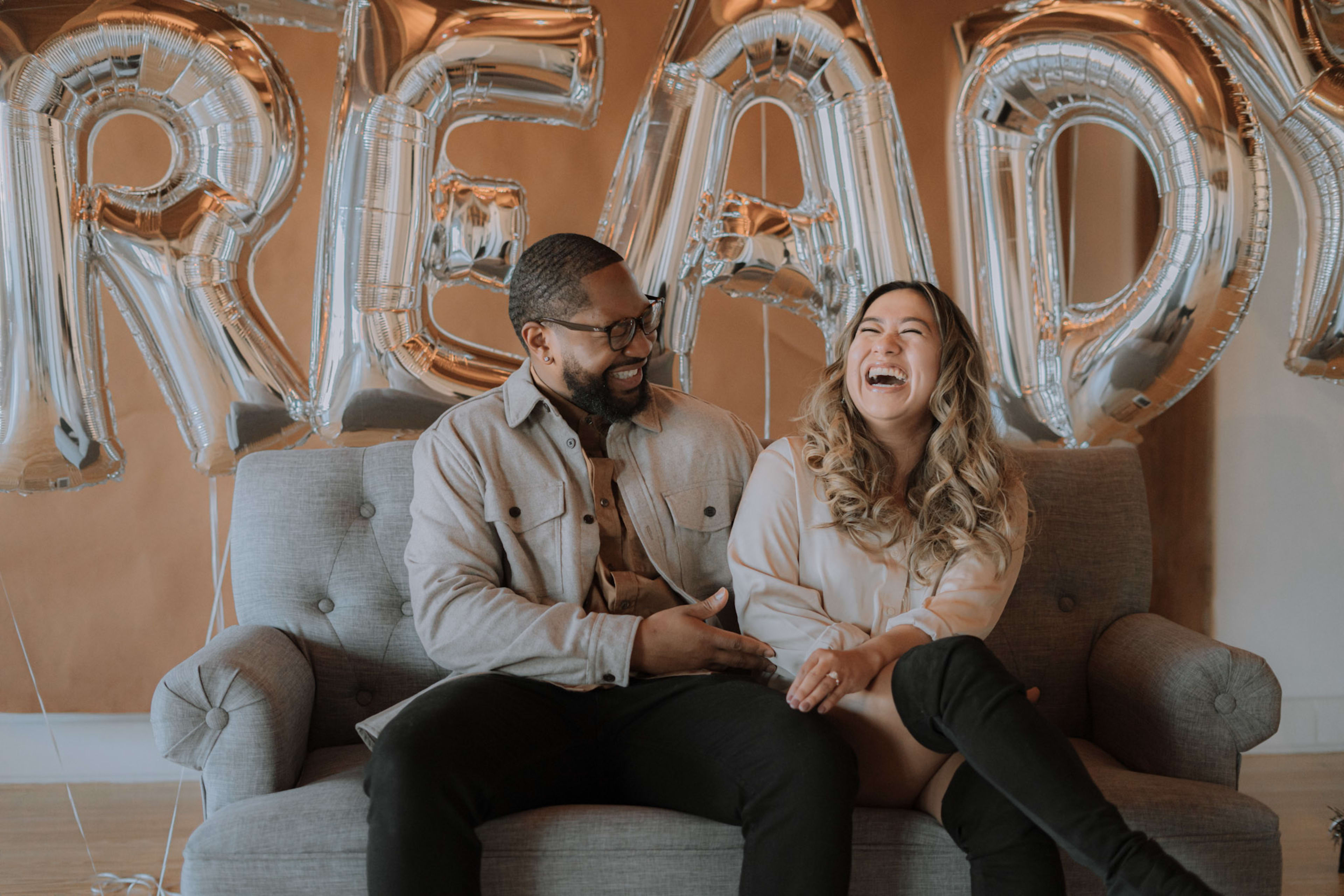 A couple on a beige couch in front of balloons at an engagement party.