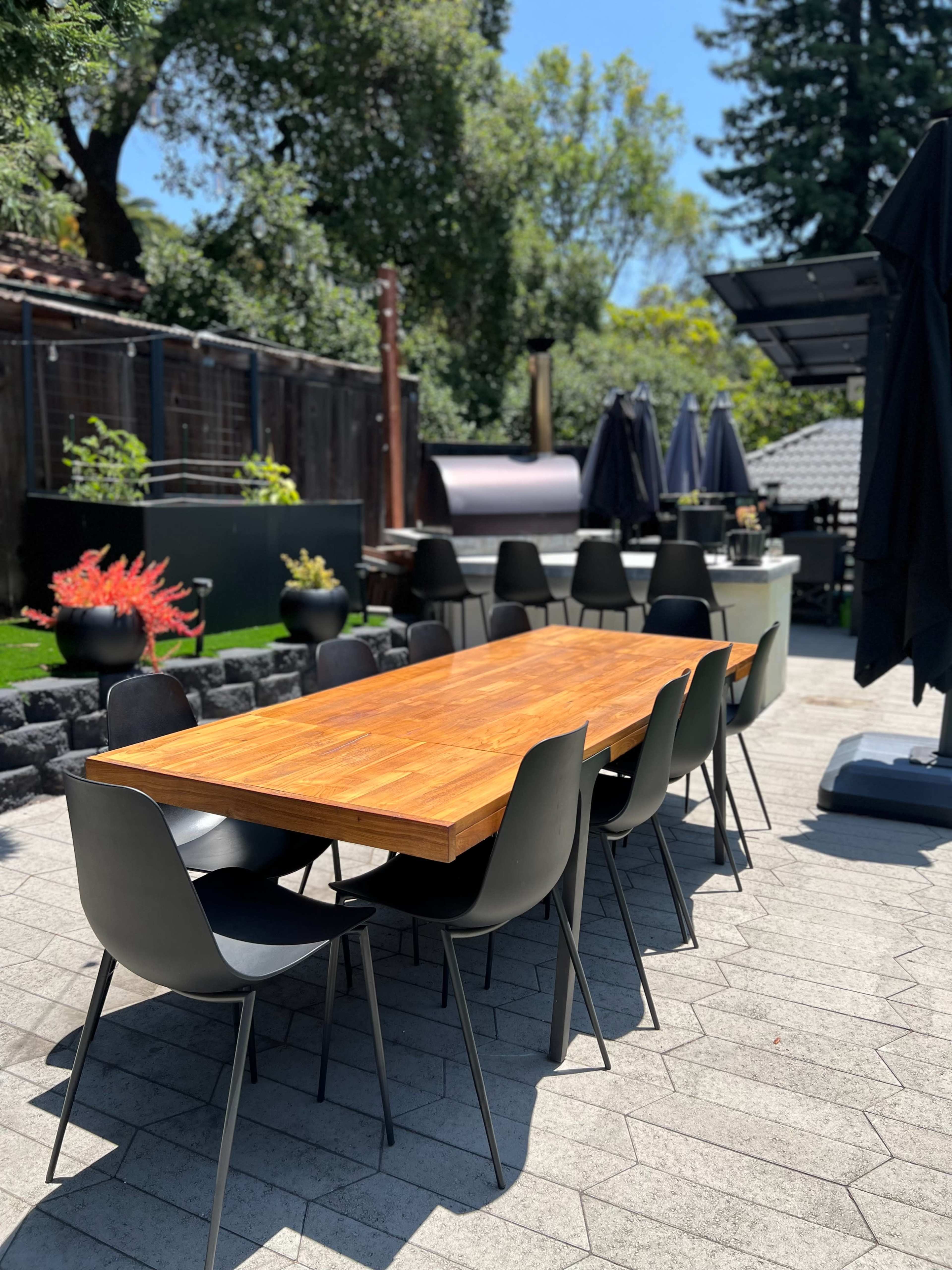 A long wooden table surrounded by black chairs is set in a sunny outdoor space with a stone patio and various plants.