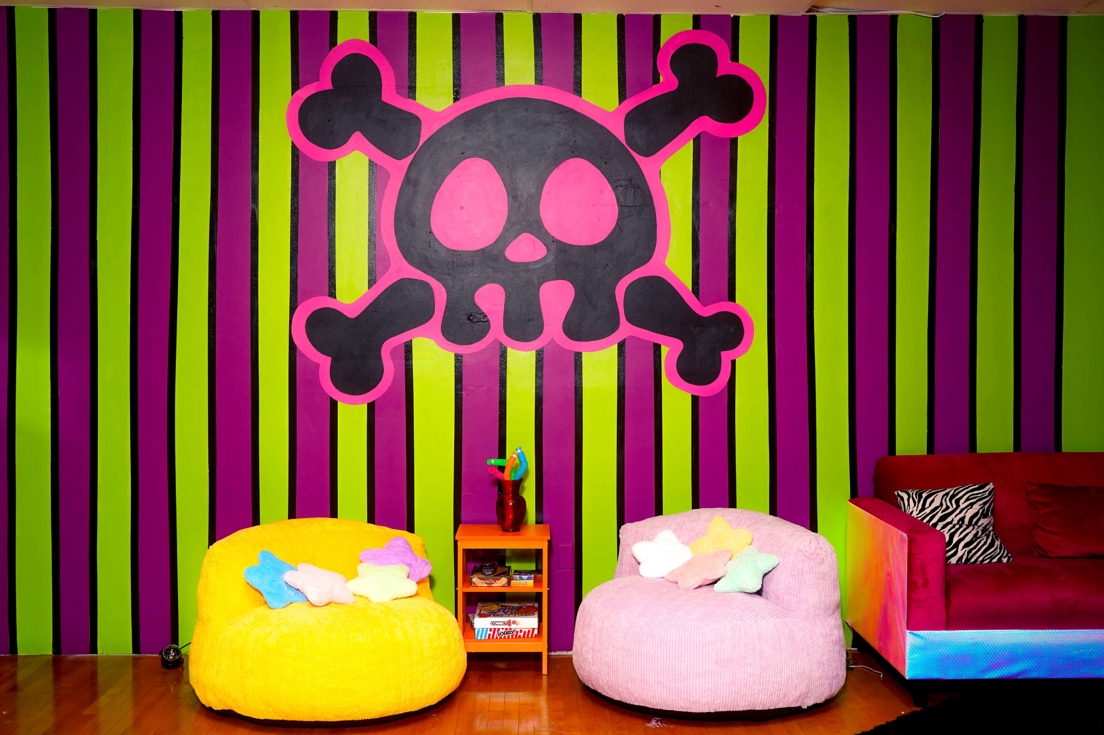 The image shows a room with bright vertical striped walls in green and black, featuring a large pink and black skull and crossbones, with two plush chairs in yellow and pink, and a small shelf holding colorful items.