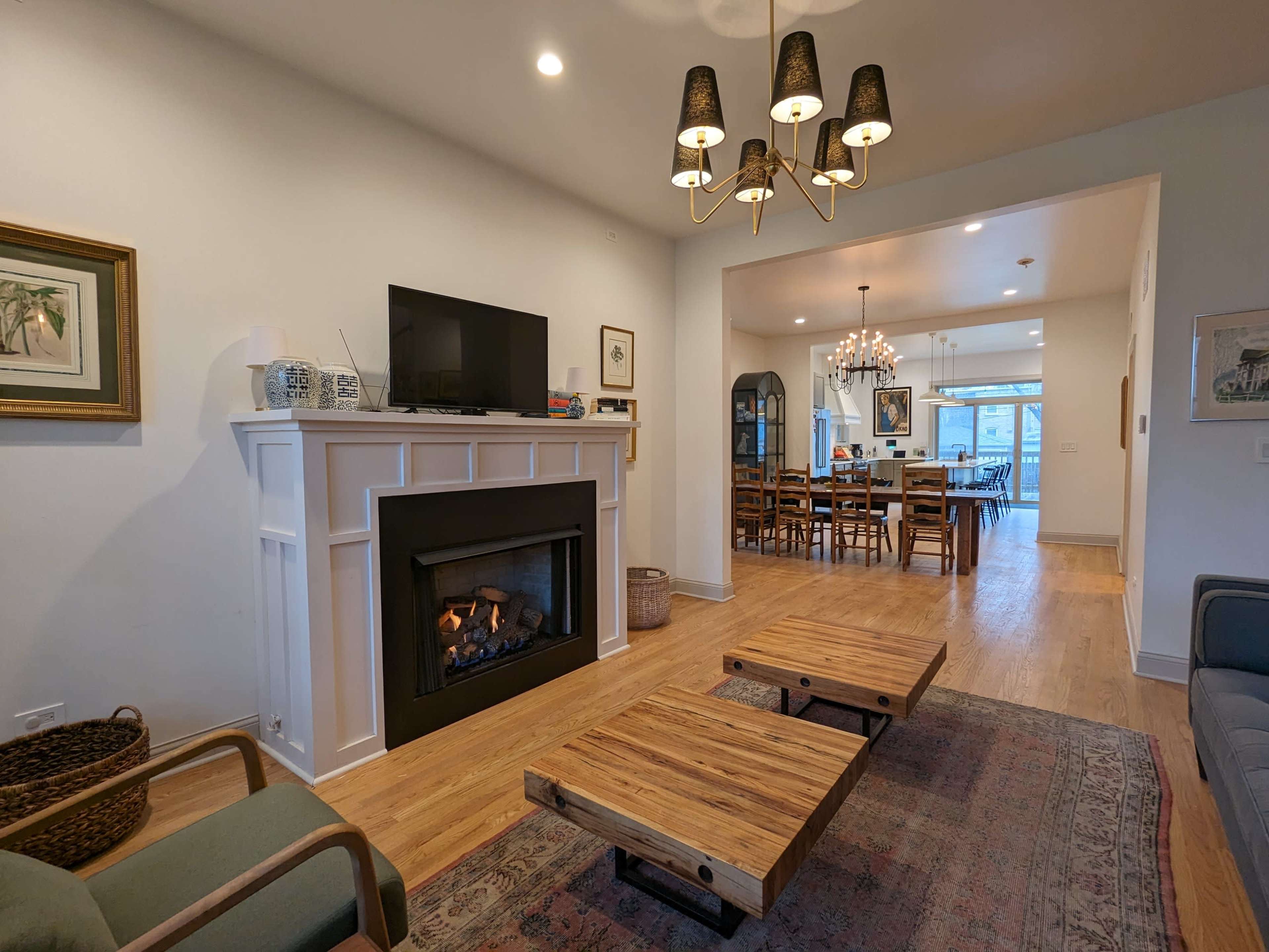 A living room featuring a modern fireplace, two wooden coffee tables, a television mounted above the mantel, and a view into a dining area with a large table.