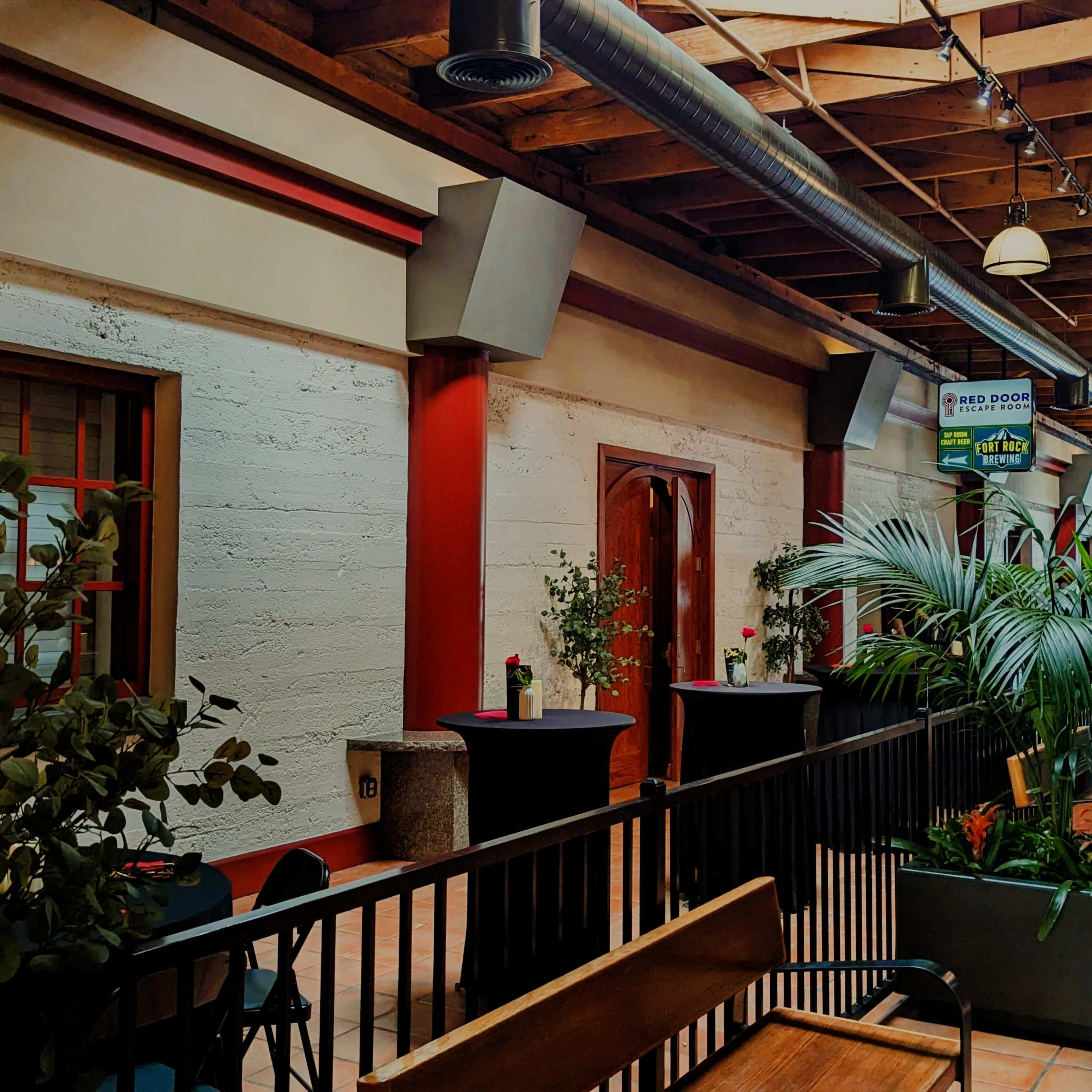 The image shows a narrow indoor space featuring a wooden ceiling, potted plants, and small round tables with floral centerpieces.