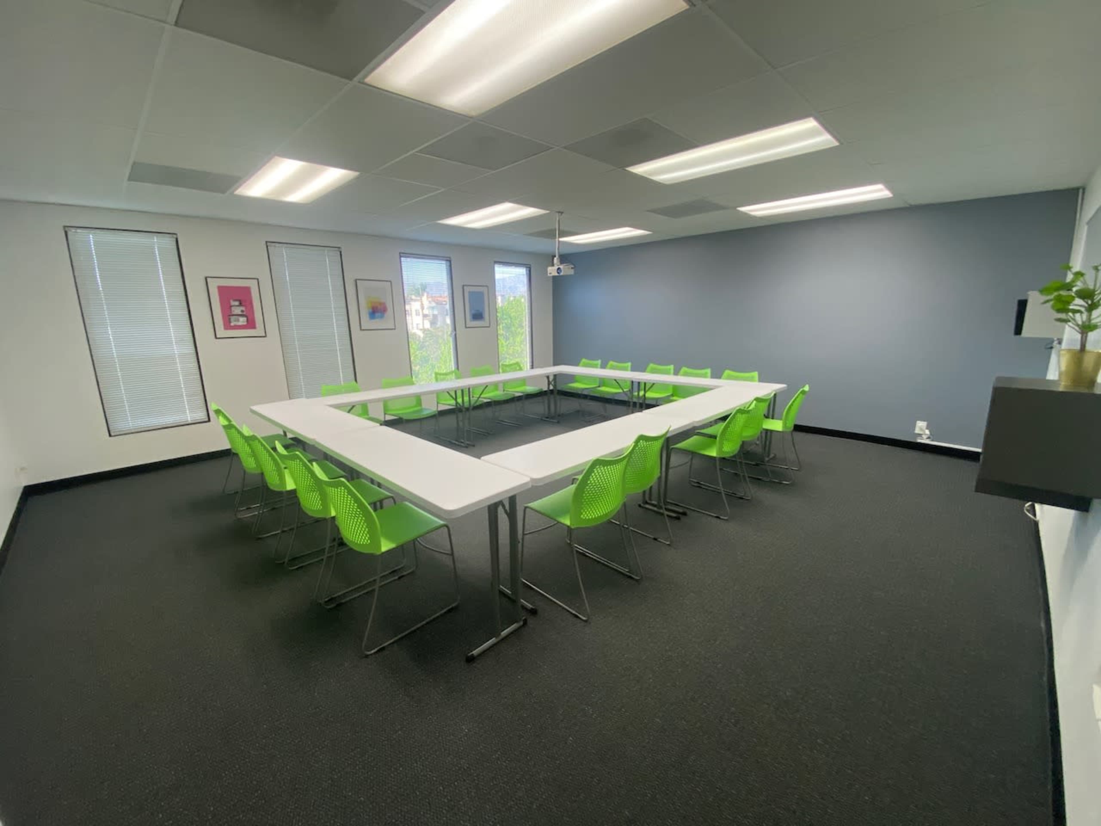 A modern meeting room features a square arrangement of white tables surrounded by green chairs, with large windows letting in natural light.