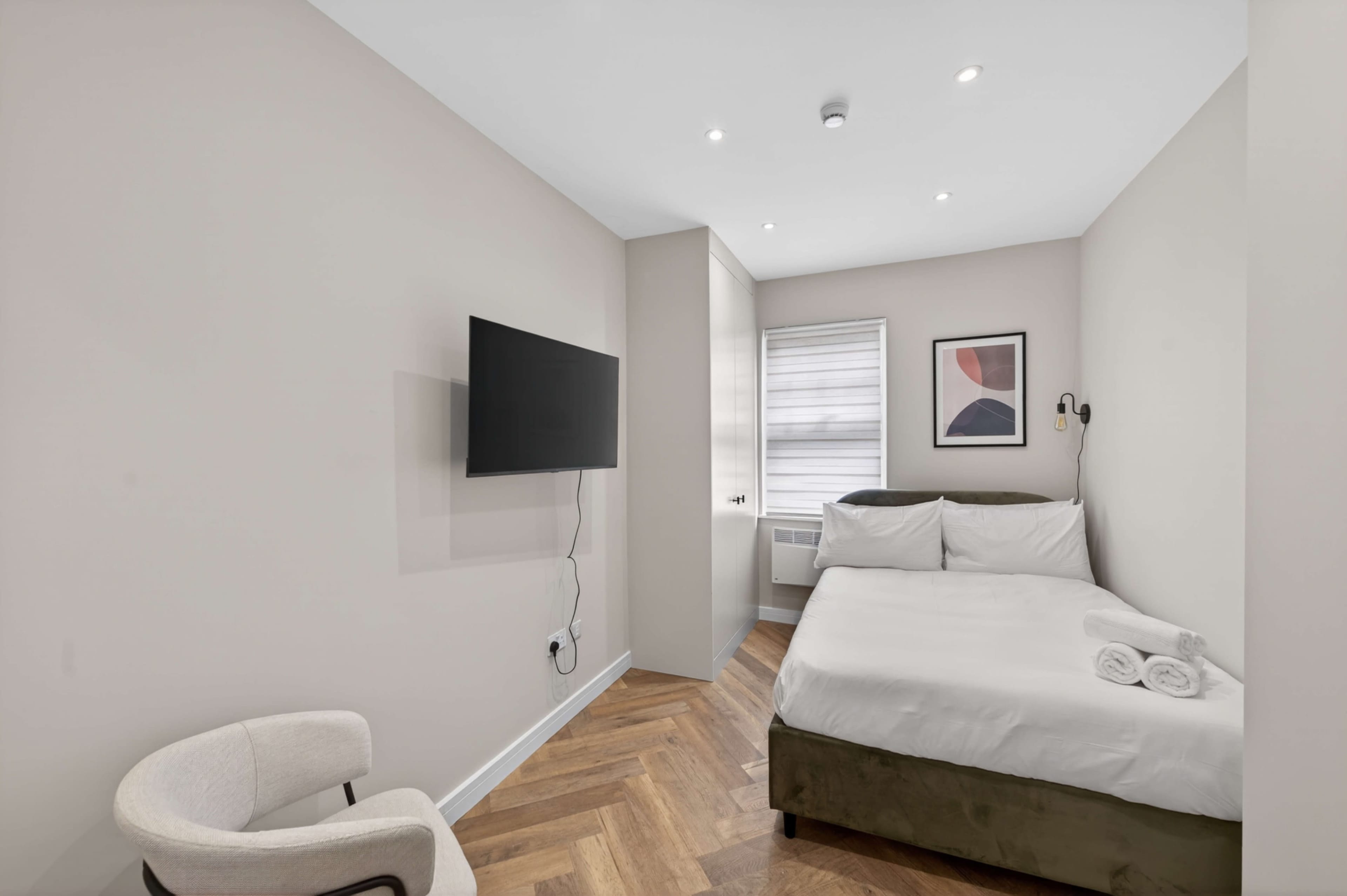 The image shows a small, modern hotel room with a bed, a television mounted on the wall, and a seating chair.