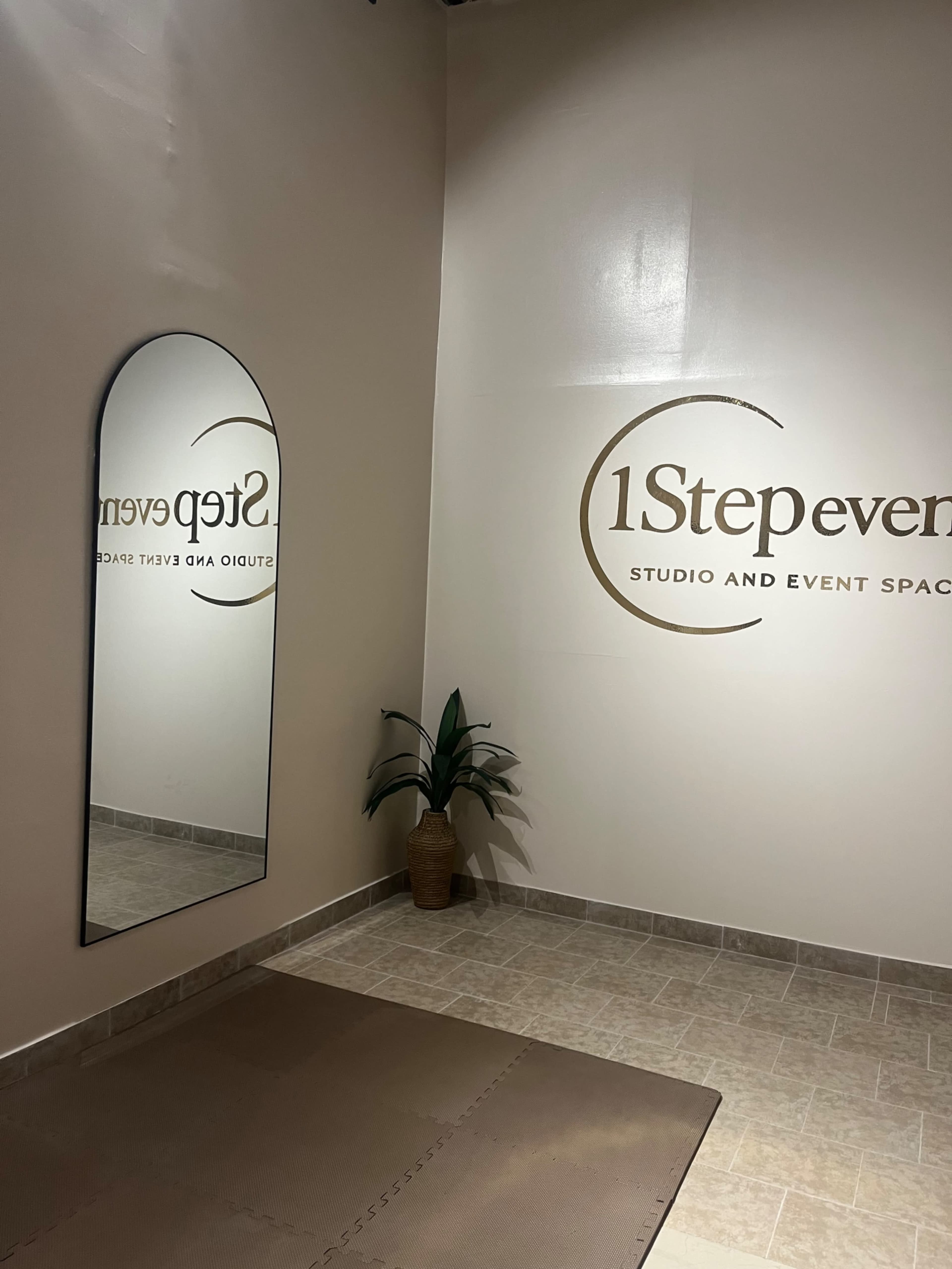 The image shows a corner of a studio or event space featuring a large mirror, a beige floor mat, and a decorative plant next to a wall with the text “1Step event studio and event space.”