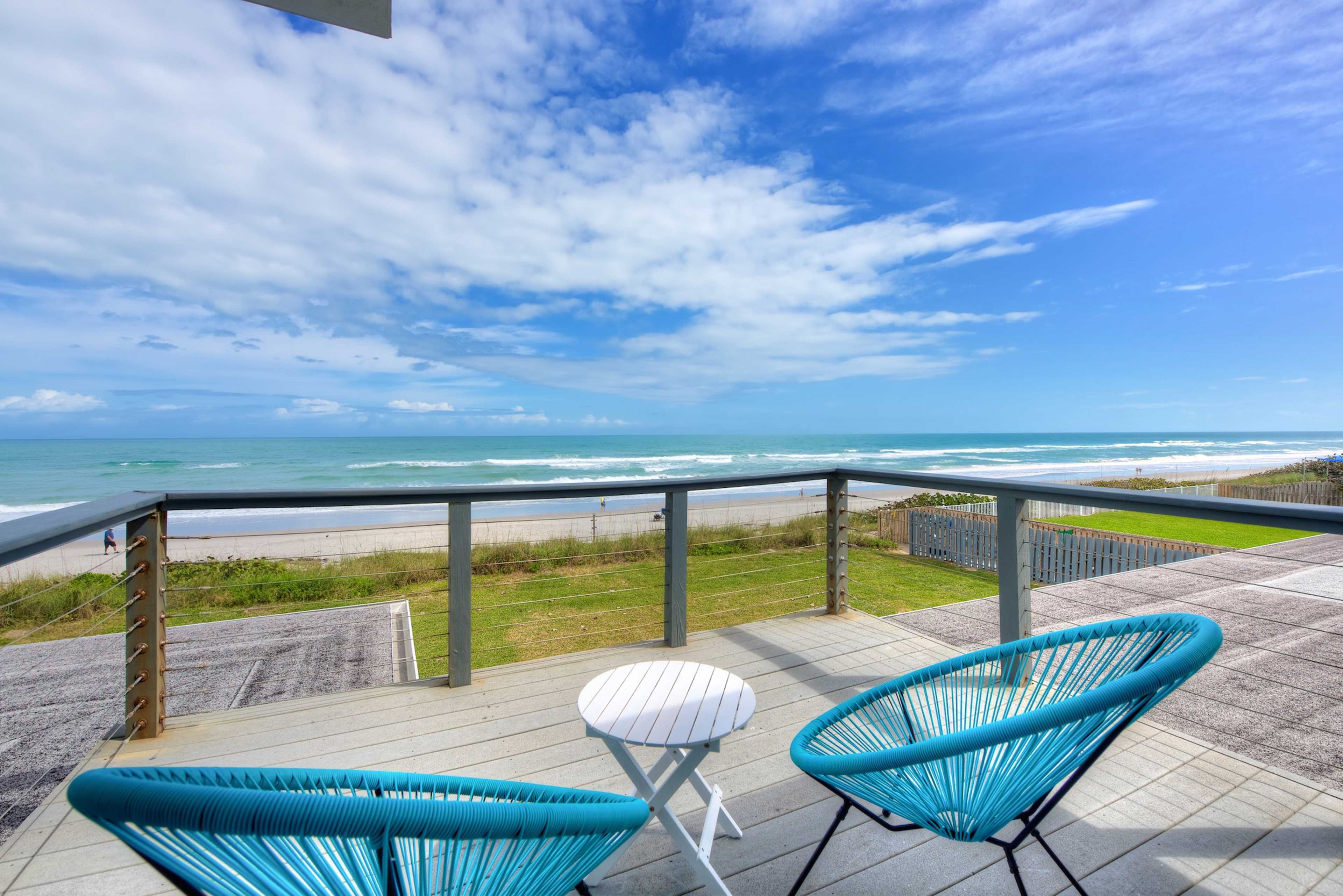 A balcony with two blue chairs overlooks a sandy beach and the ocean under a partly cloudy sky.