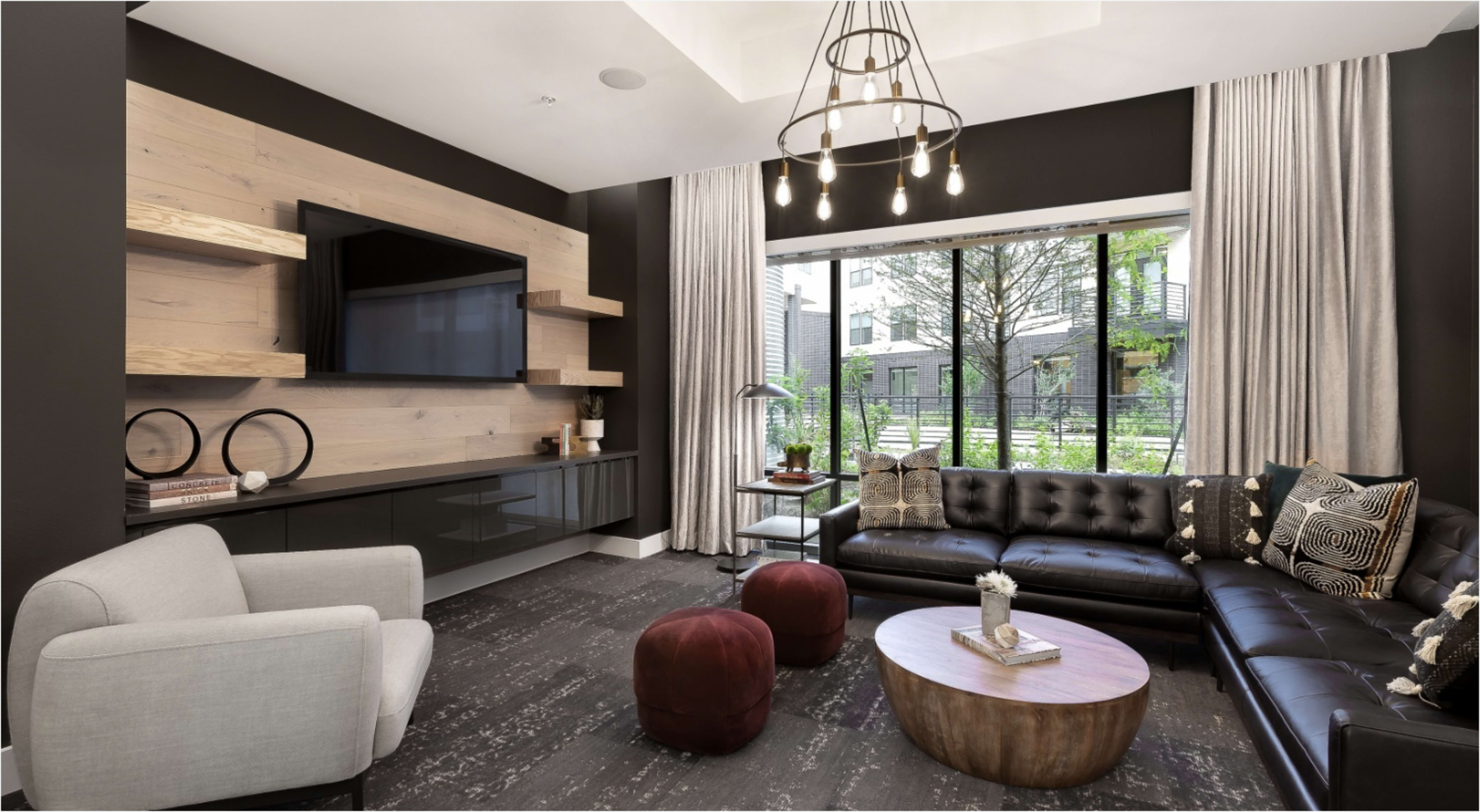 The image shows a modern living room featuring a large black leather sectional sofa, a wooden coffee table, and a wall-mounted television, with large windows overlooking a green outdoor area.