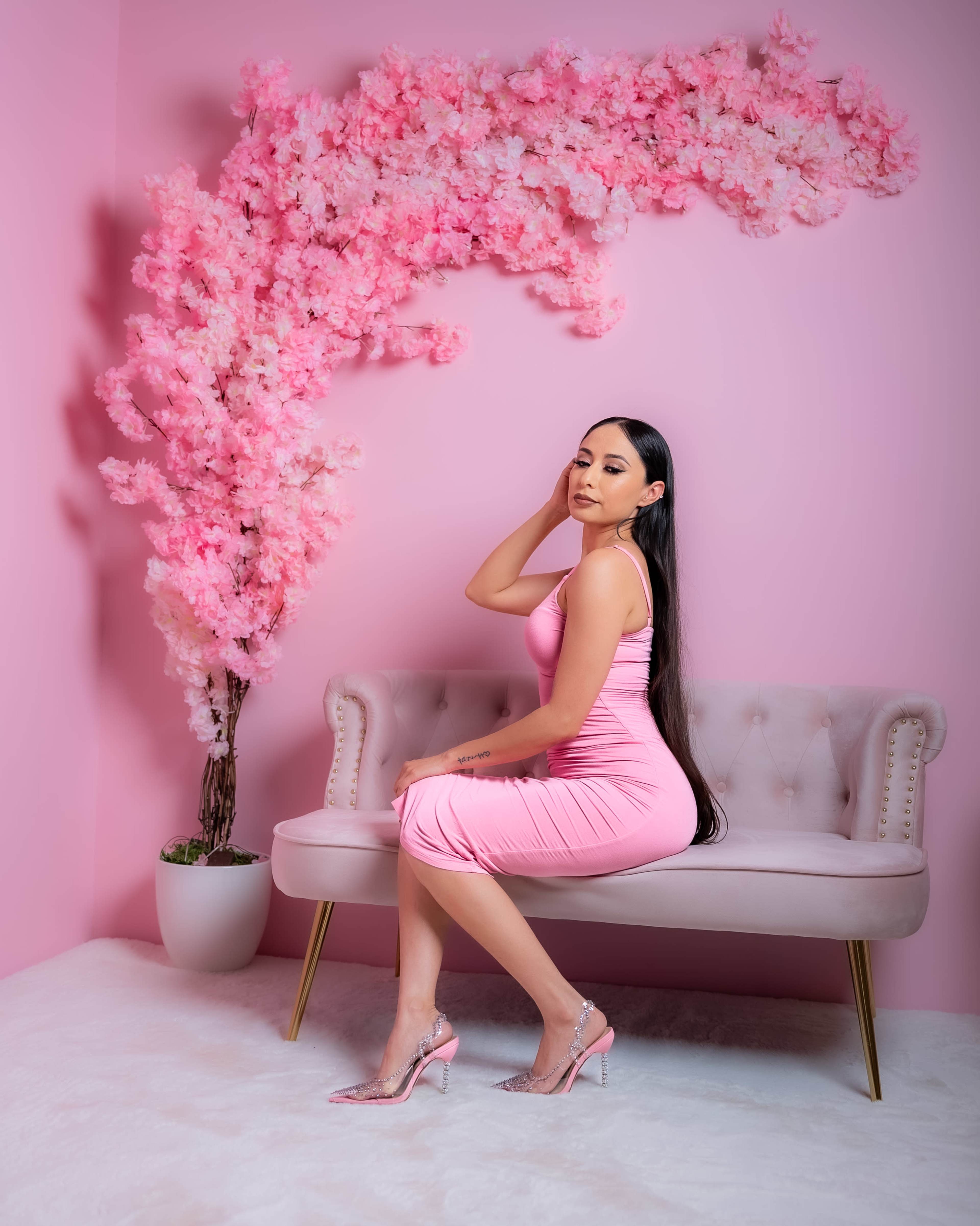 A woman in a pink dress sits on a light-colored velvet sofa against a backdrop of pink floral decorations.