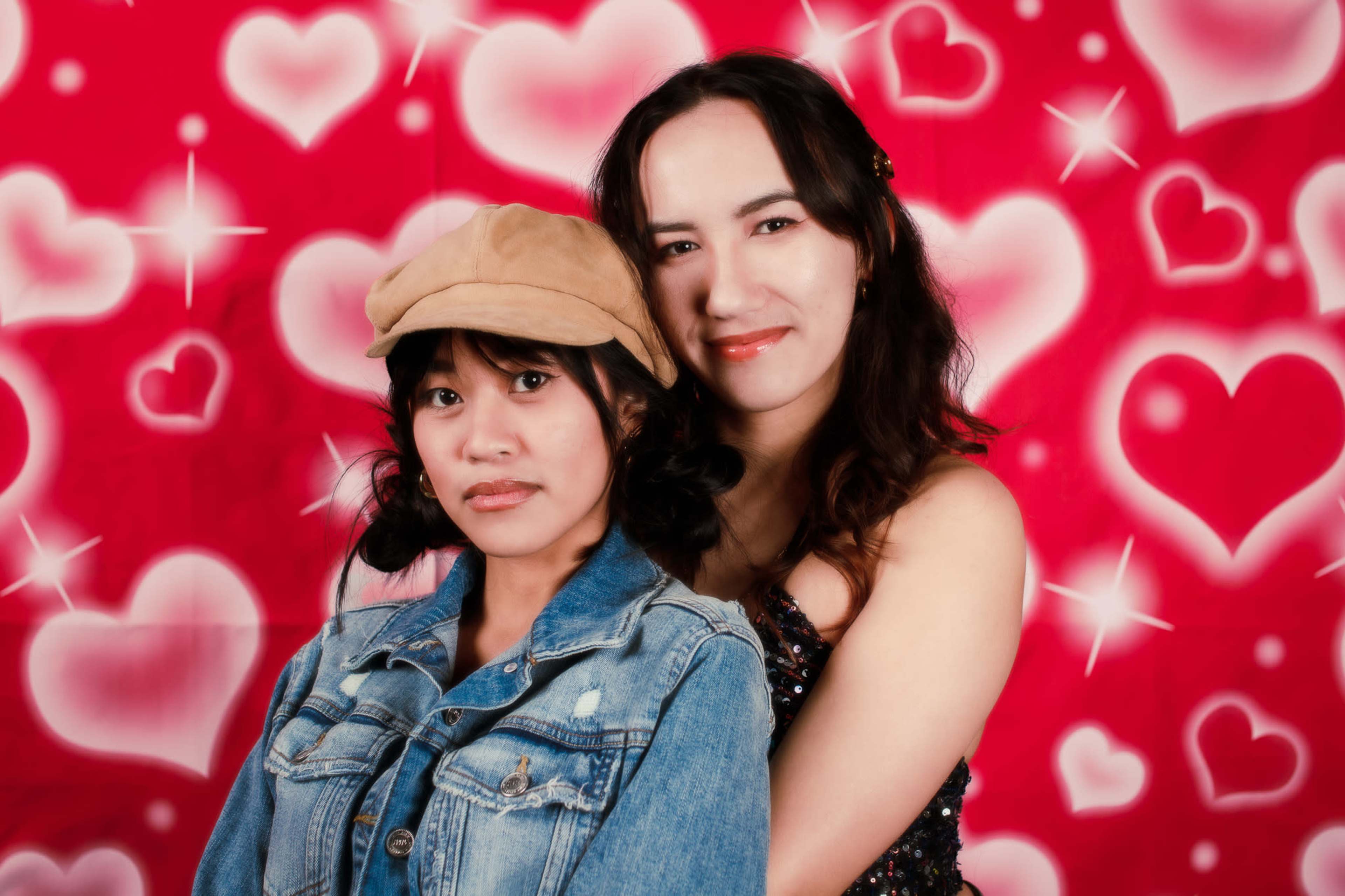 Two young women pose together in front of a red backdrop decorated with hearts.