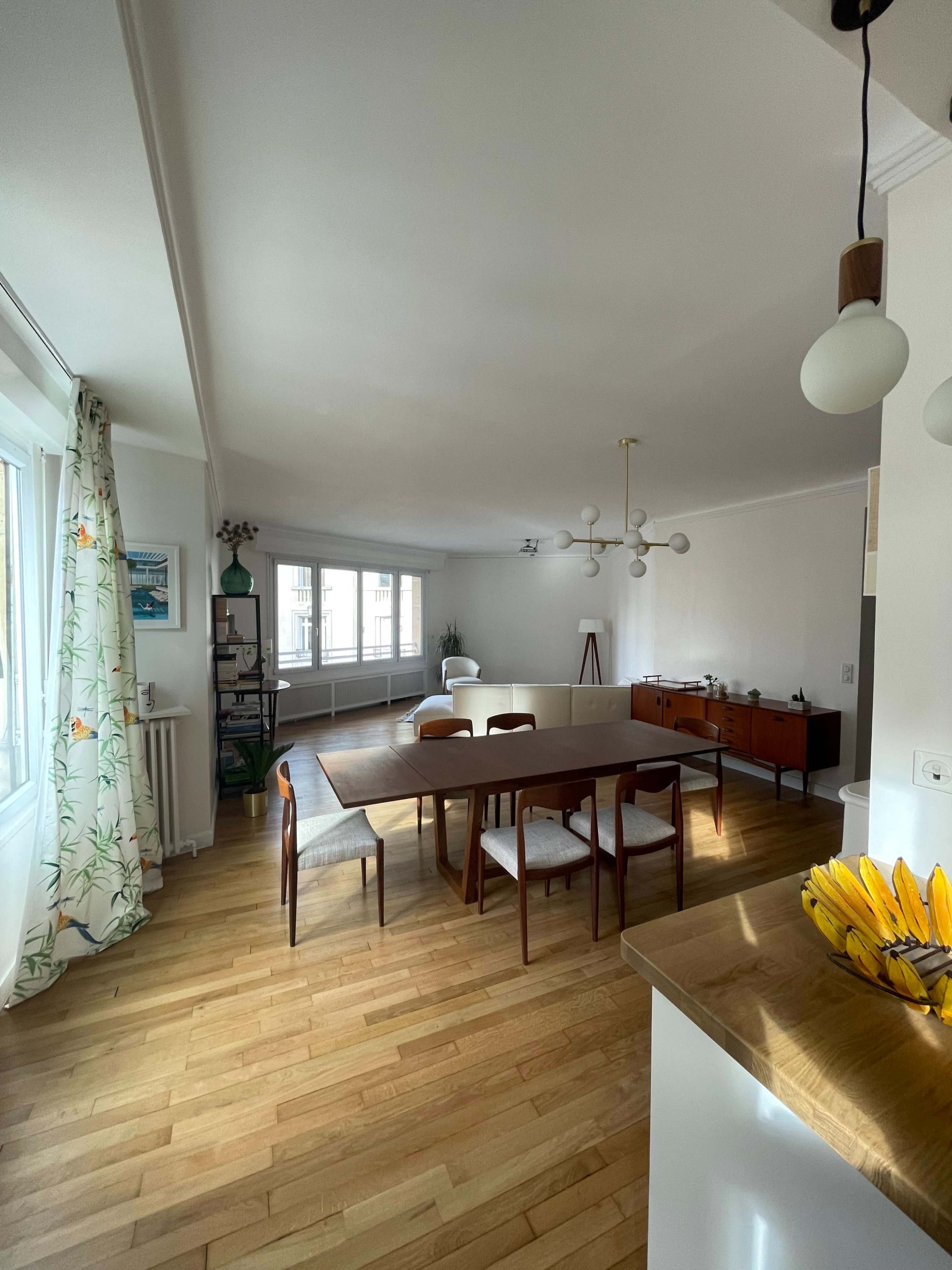 A modern dining area features a wooden table surrounded by minimalist chairs, with large windows allowing natural light and a sideboard in the background.