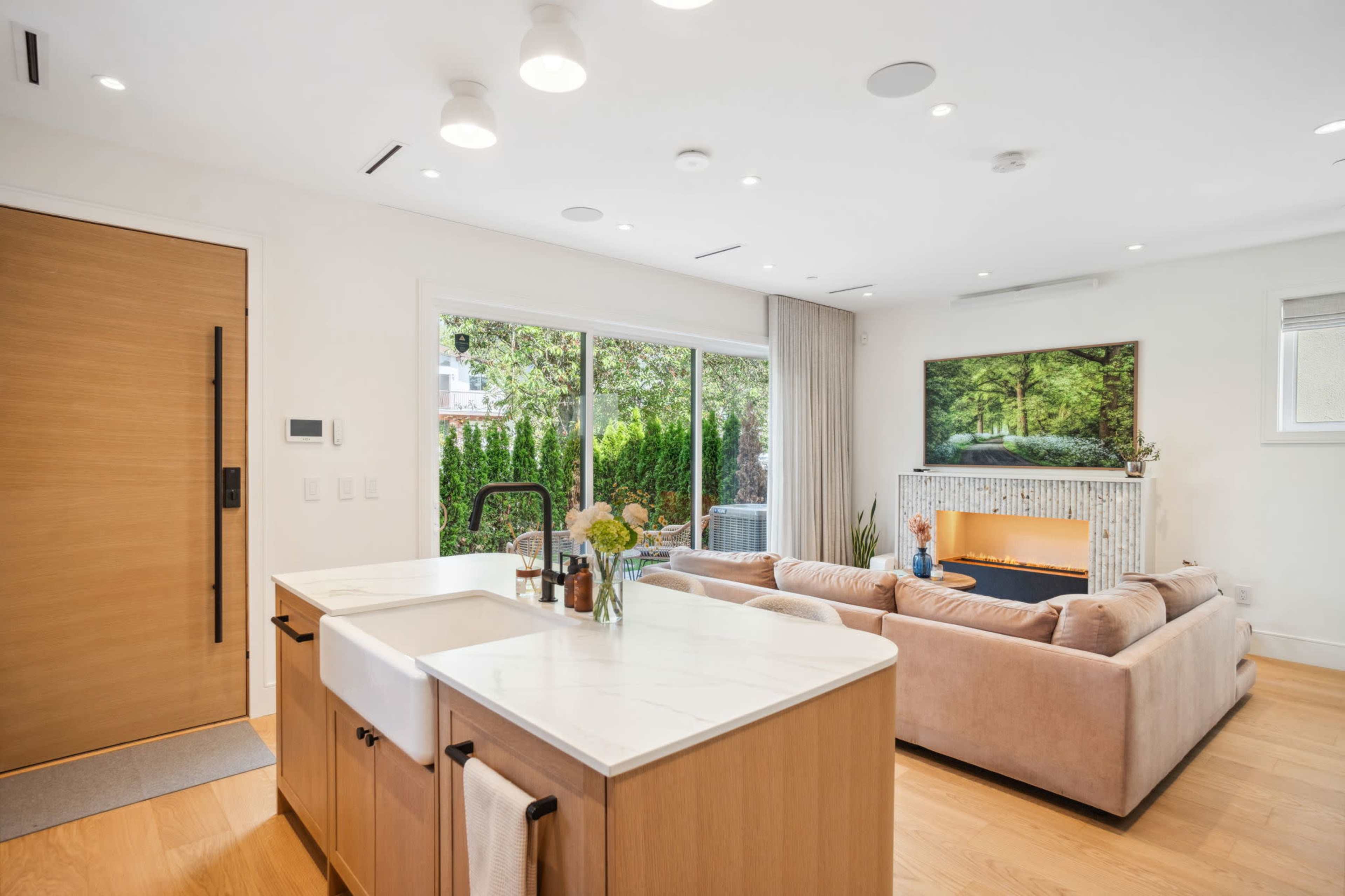A modern open-concept living space features a kitchen island with a sink, a cozy seating area with a sofa, and large windows overlooking a green outdoor area.