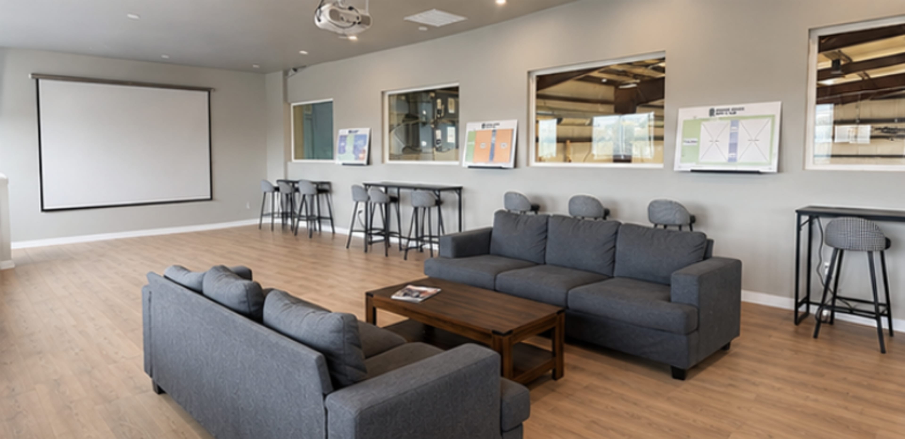 The image shows a modern lounge area with two gray sofas, a wooden coffee table, a projector, and several high chairs positioned near large windows.