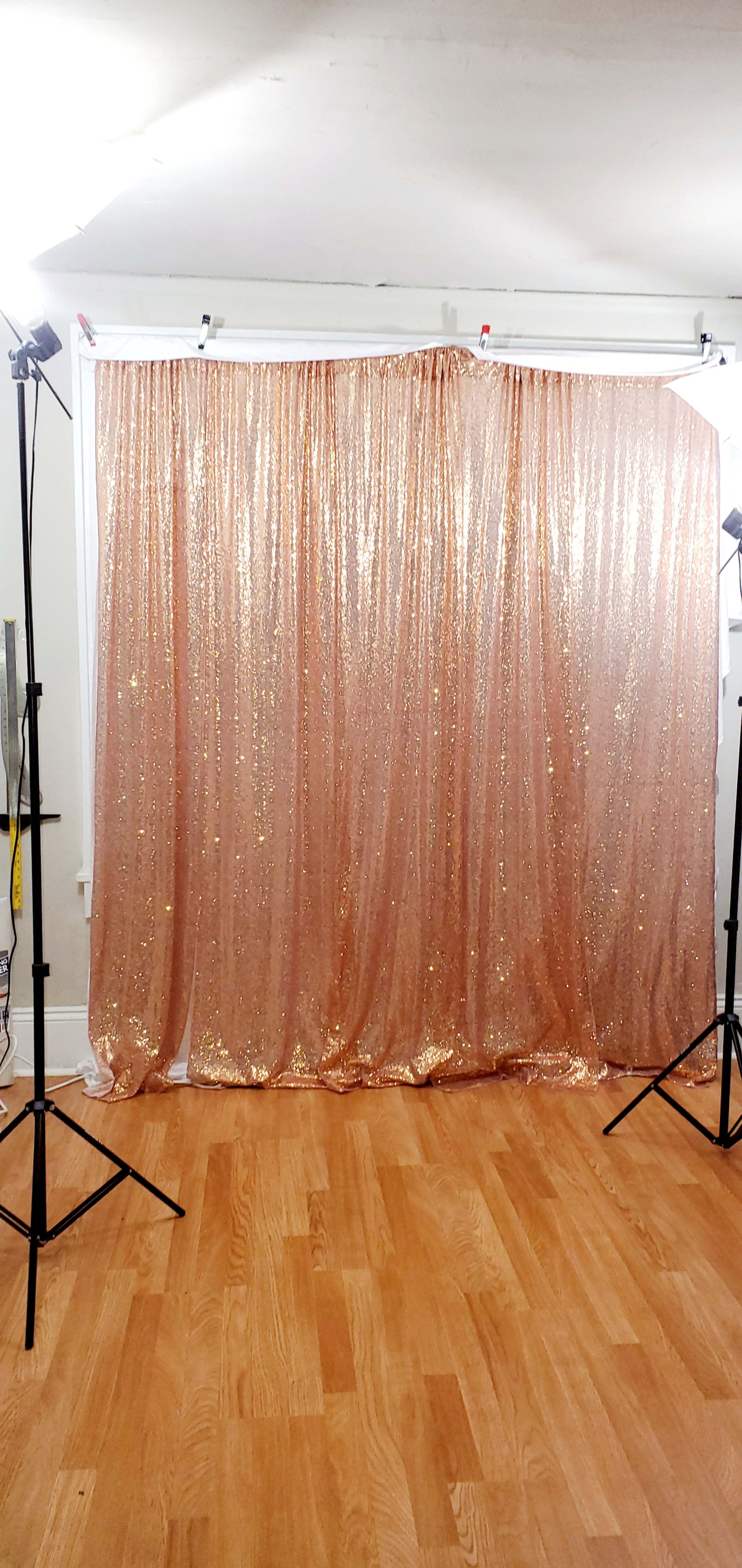 A backdrop of shimmering rose gold sequined fabric set up in a room with wooden flooring, accompanied by two standing lights on tripods.