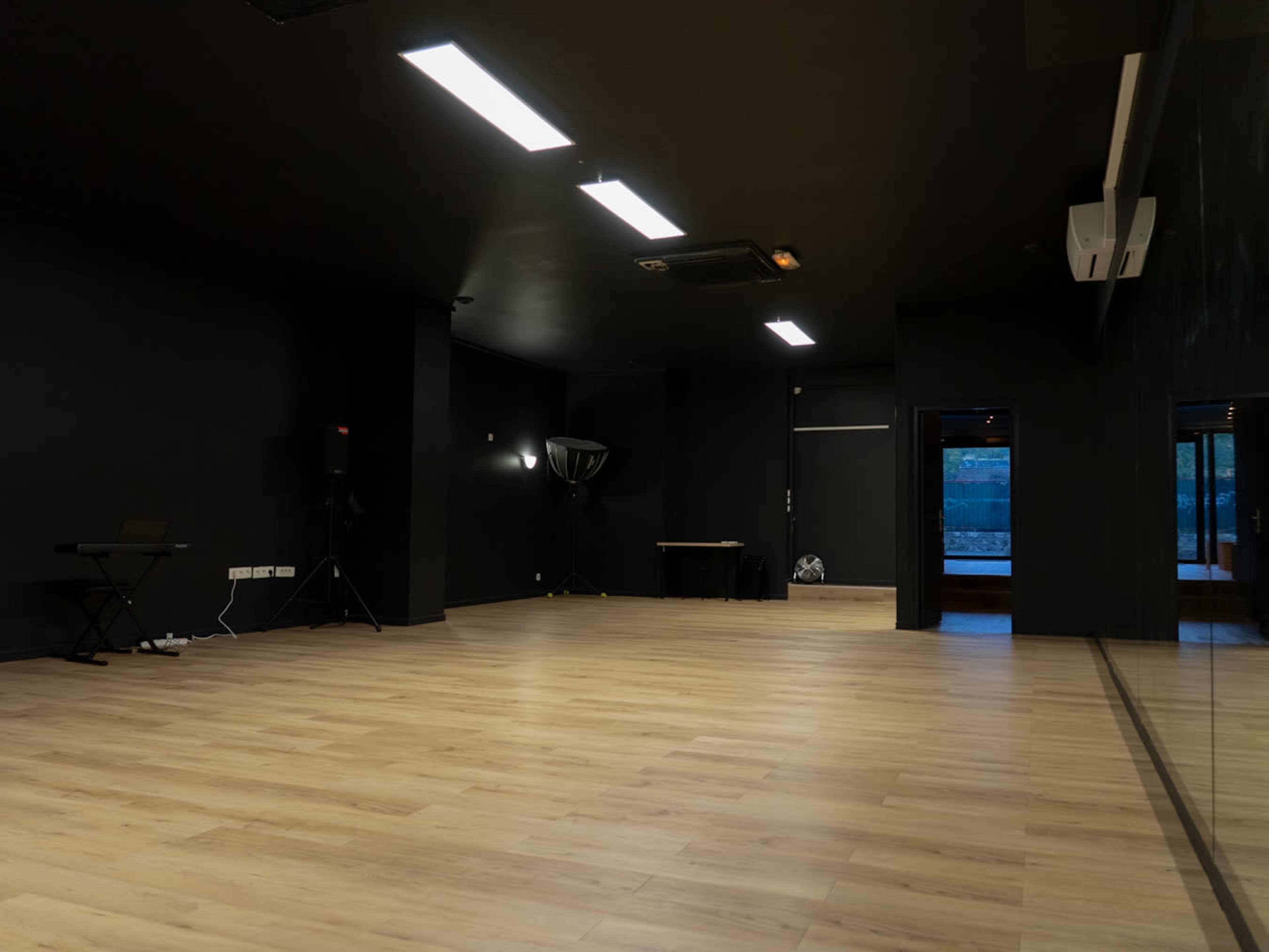 The image shows a spacious, empty room with a wooden floor and black walls, illuminated by overhead lighting.