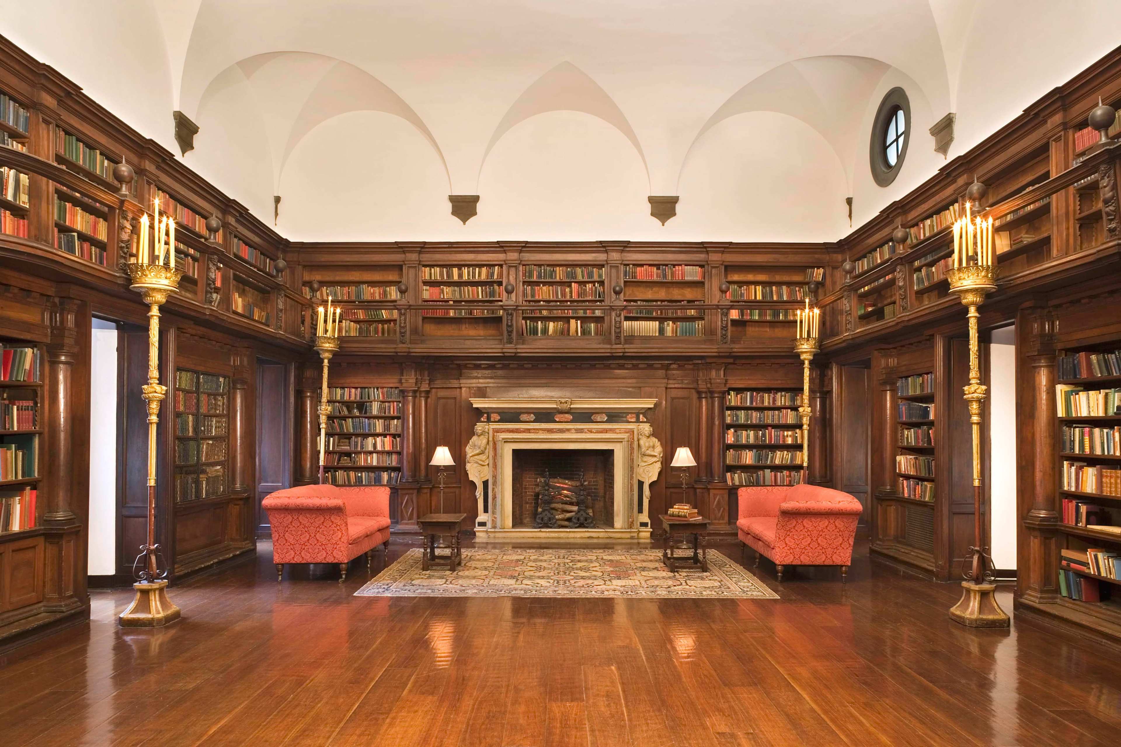 The image shows a spacious library with wooden bookshelves filled with books, two red upholstered sofas, and a fireplace at the center.
