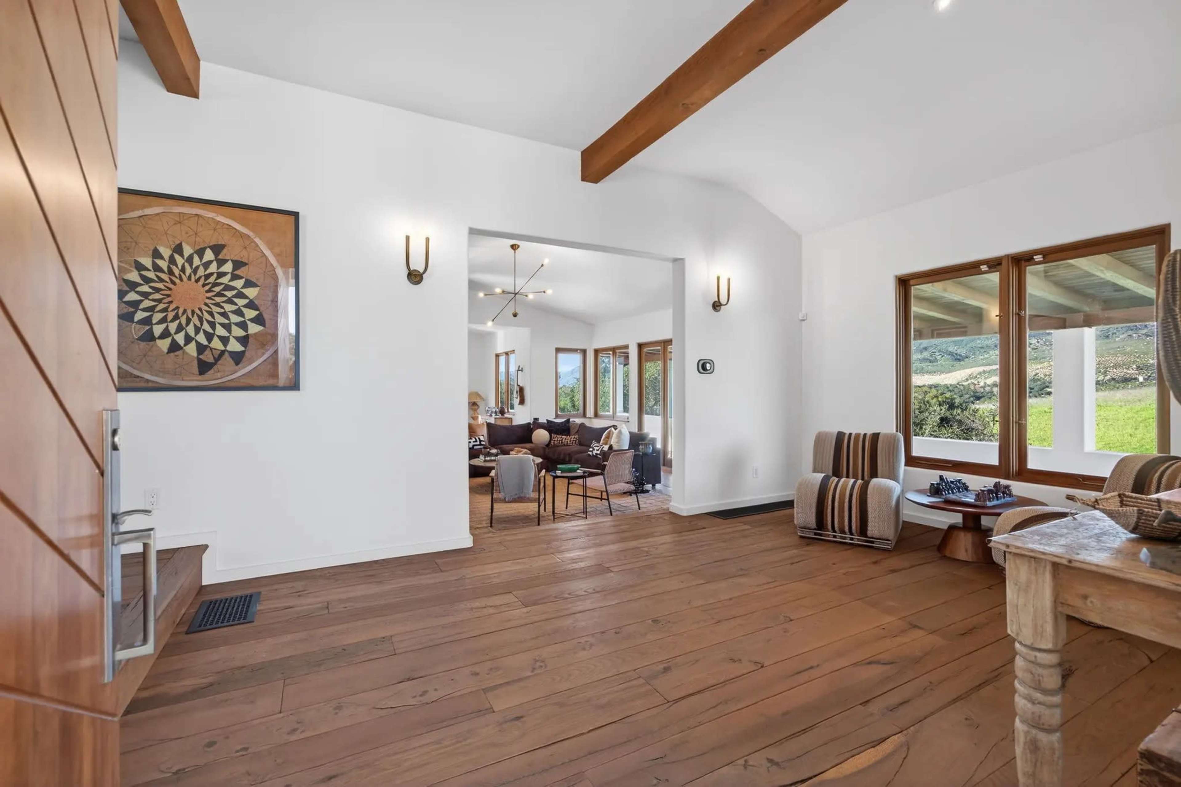 The image shows a bright living space with wooden flooring, exposed beams, and large windows that open to a view of green fields.