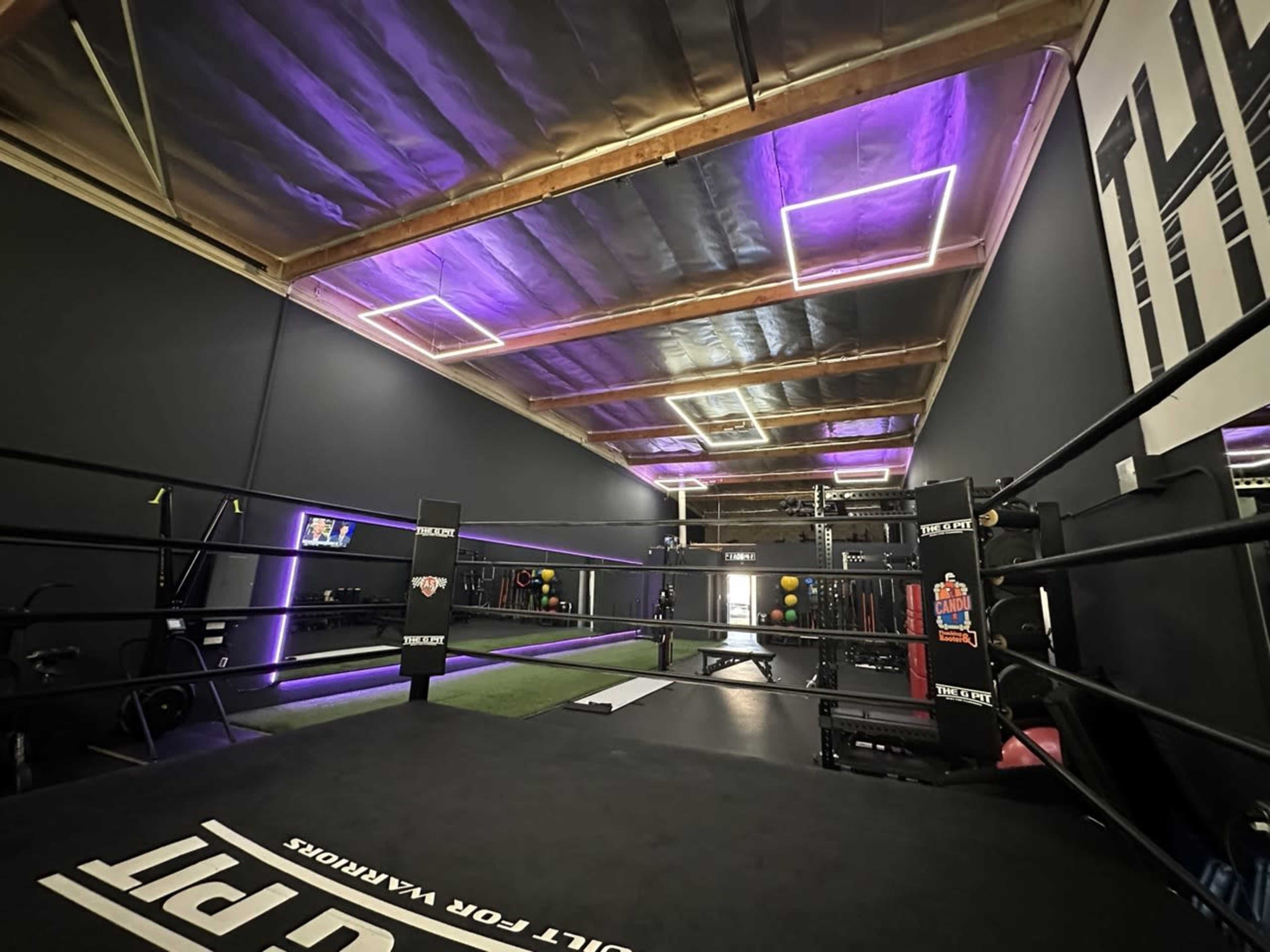 The image shows a modern boxing gym with a ring at the center, illuminated by purple LED lights and equipped with various training equipment along the walls.