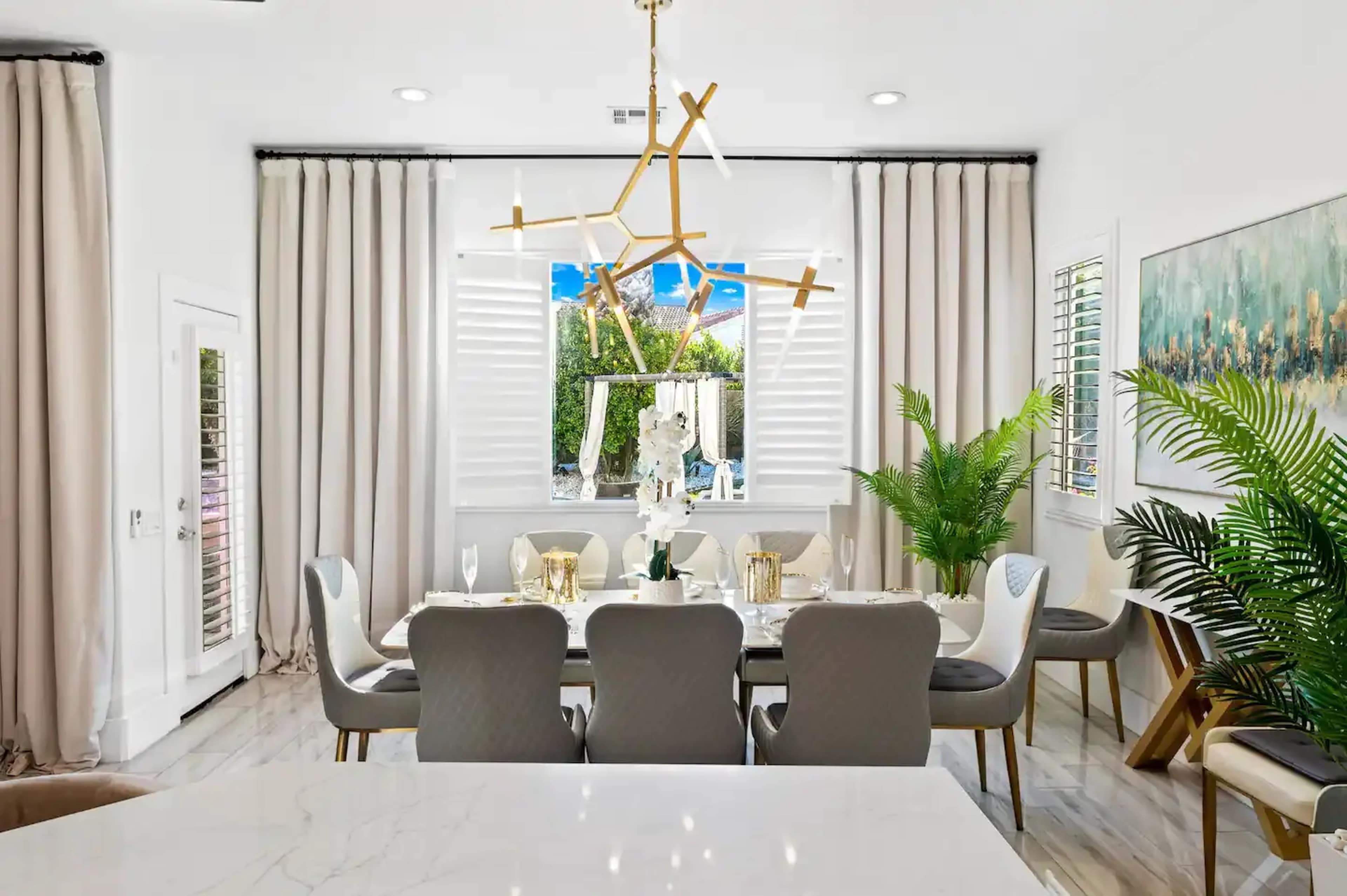 A dining room features a large table set for meals, surrounded by chairs, with a modern light fixture and potted plants near the windows.
