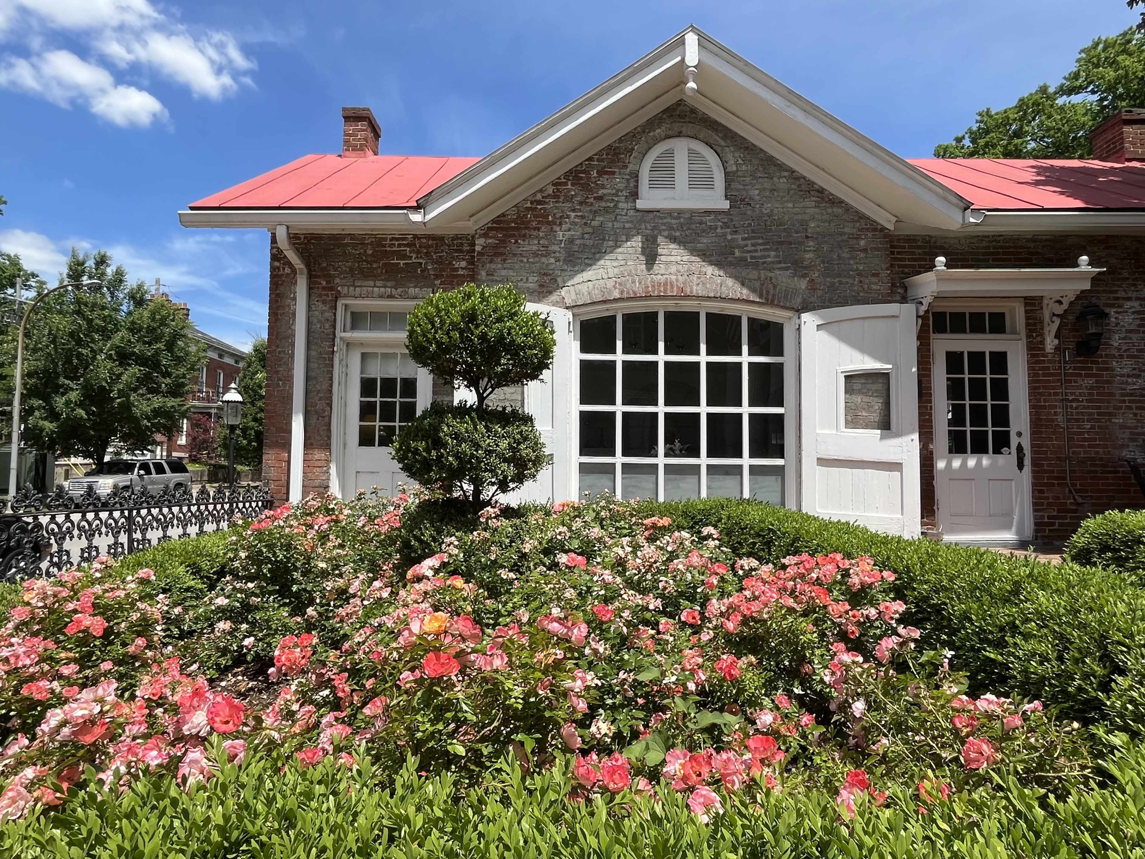 A brick house with a red metal roof features large windows and is surrounded by a blooming flower bed filled with pink and orange flowers.