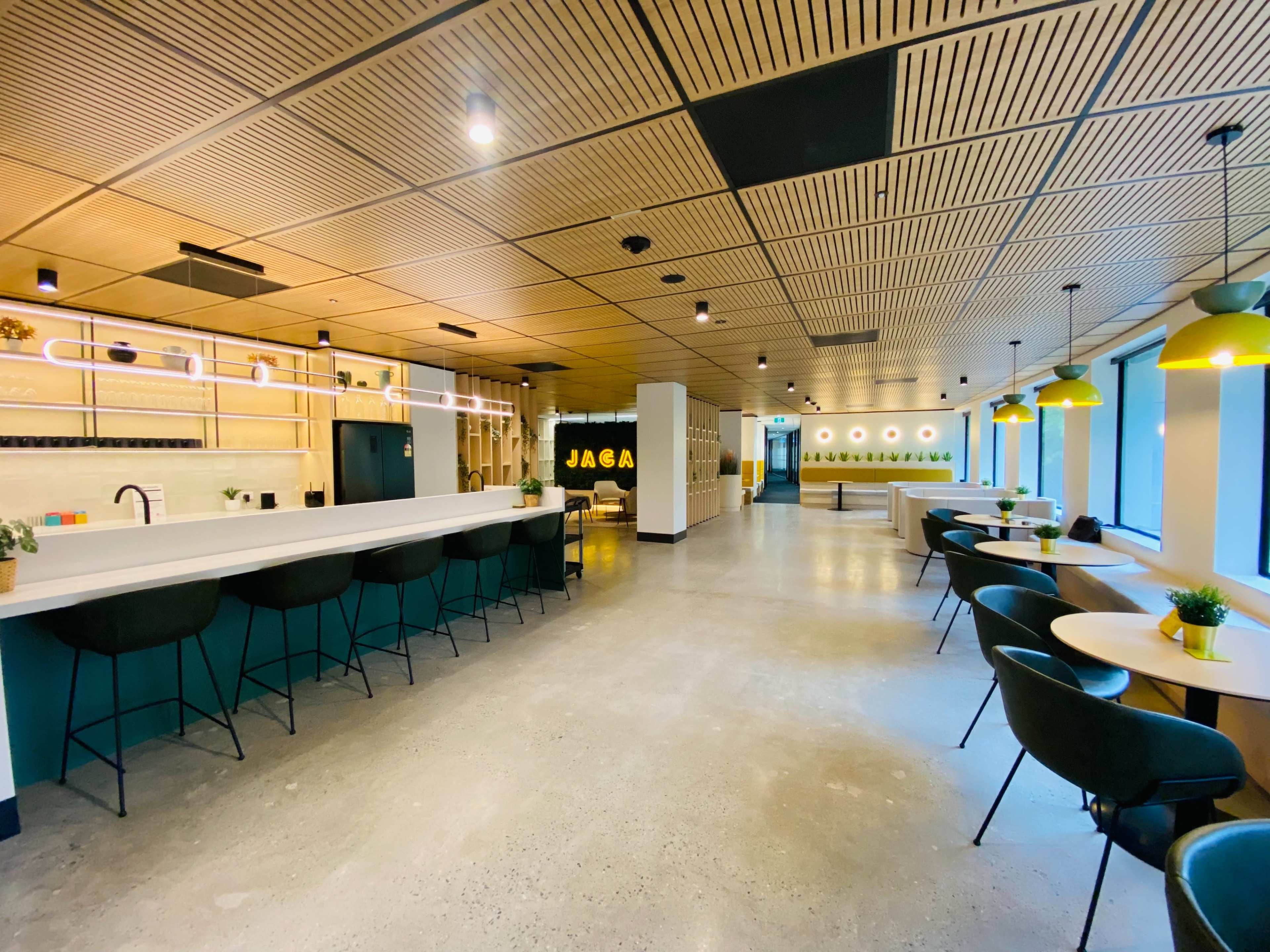 The image shows a modern office space with a long counter, seating areas, and a green accent wall, all featuring large windows and contemporary lighting.